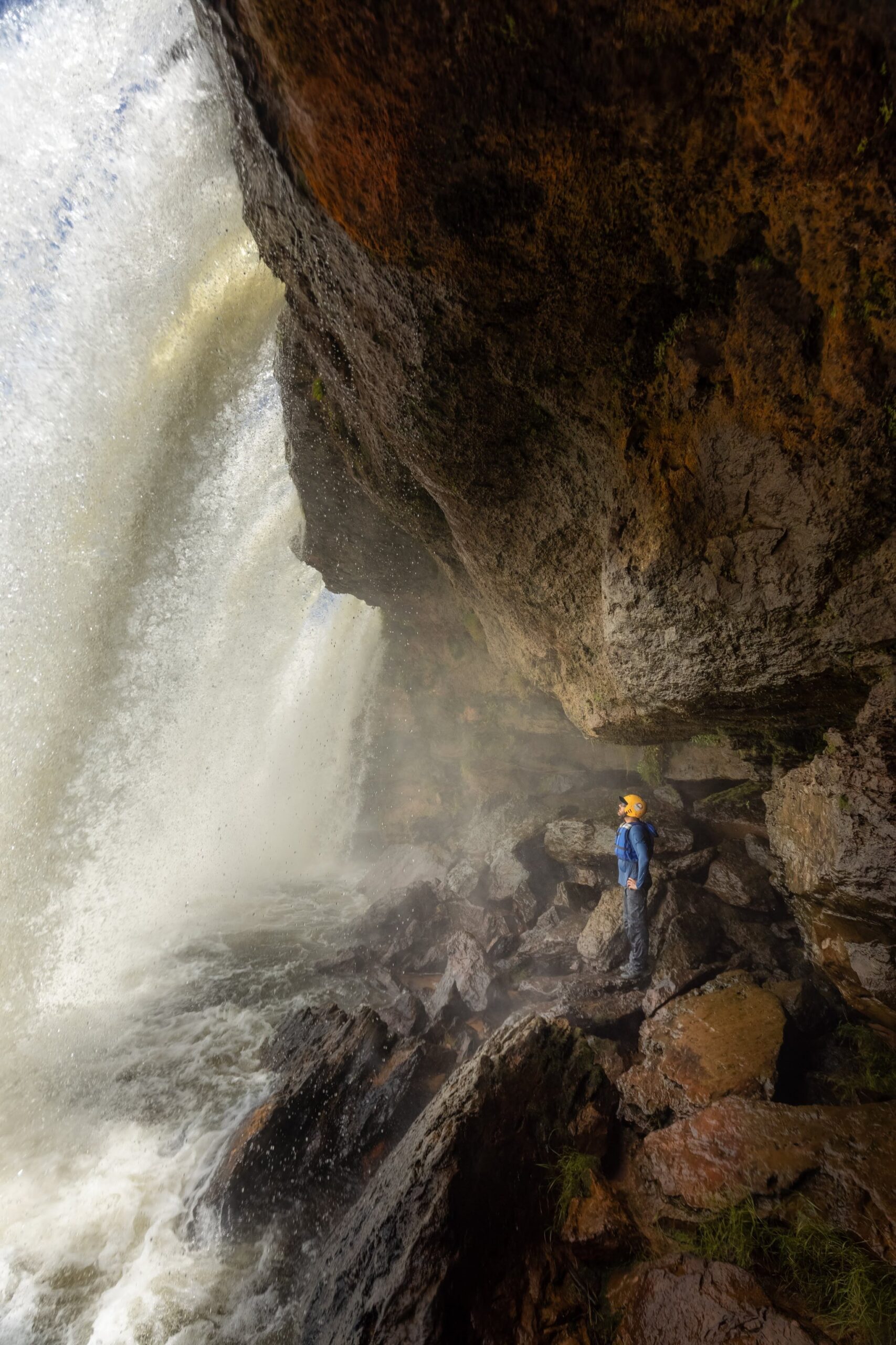 Verpletterd door mist en geraas — de waterval maakt de mens klein. Photo: Alexandre Socci Een avonturier met helm en blauwe jas staat op een rotsrichel onder een donderende waterval tijdens een Jalapão-avontuur, volledig overheerst door de overhangende klif en wervelende nevel