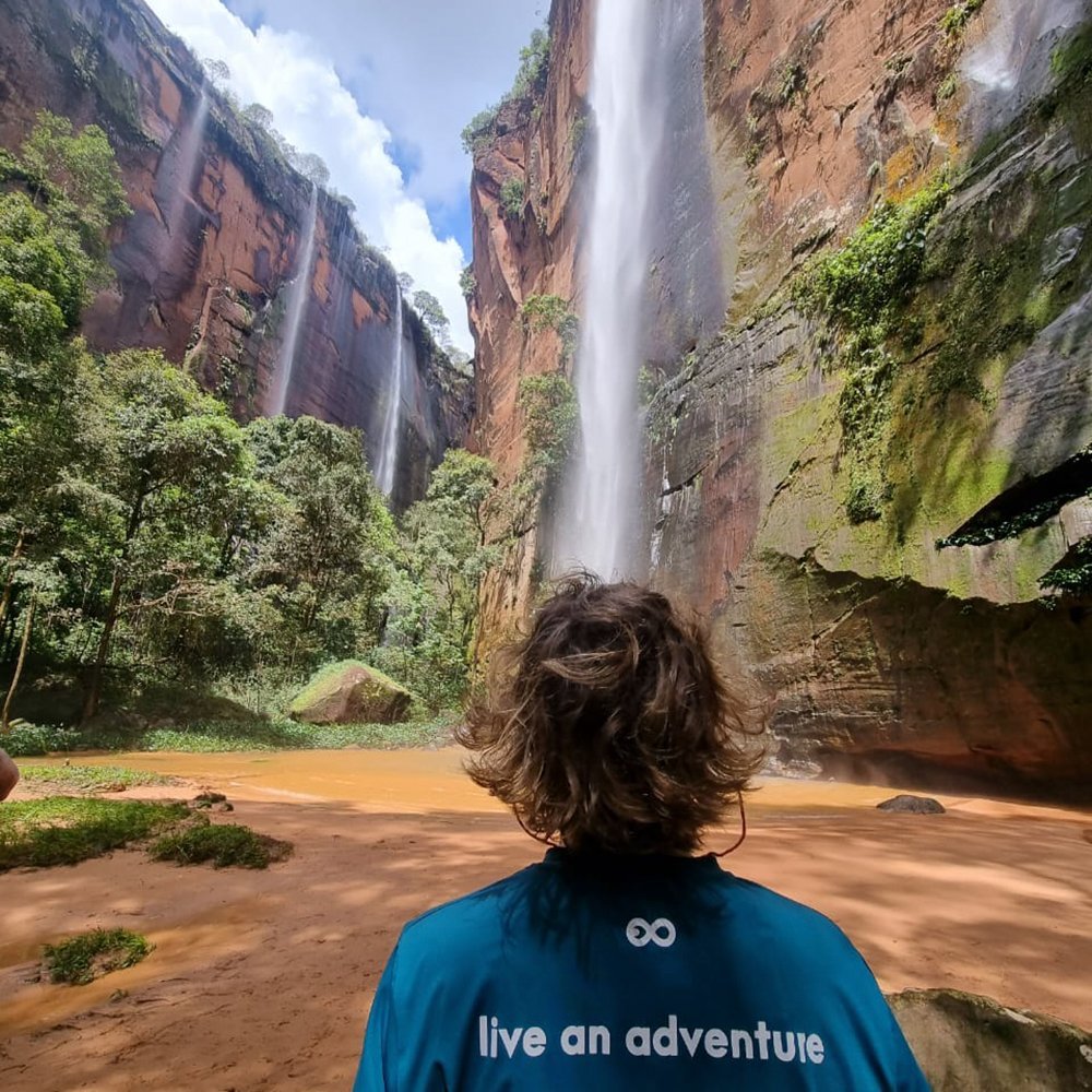 Meerdere watervallen tegelijk — rood zandsteen op zijn indrukwekkendst. Een reiziger in een blauwgroen shirt staart omhoog naar meerdere watervallen die langs rode zandsteenkliffen neerstorten in een okerkleurig bassin — een hoogtepunt van het Jalapão-avontuur