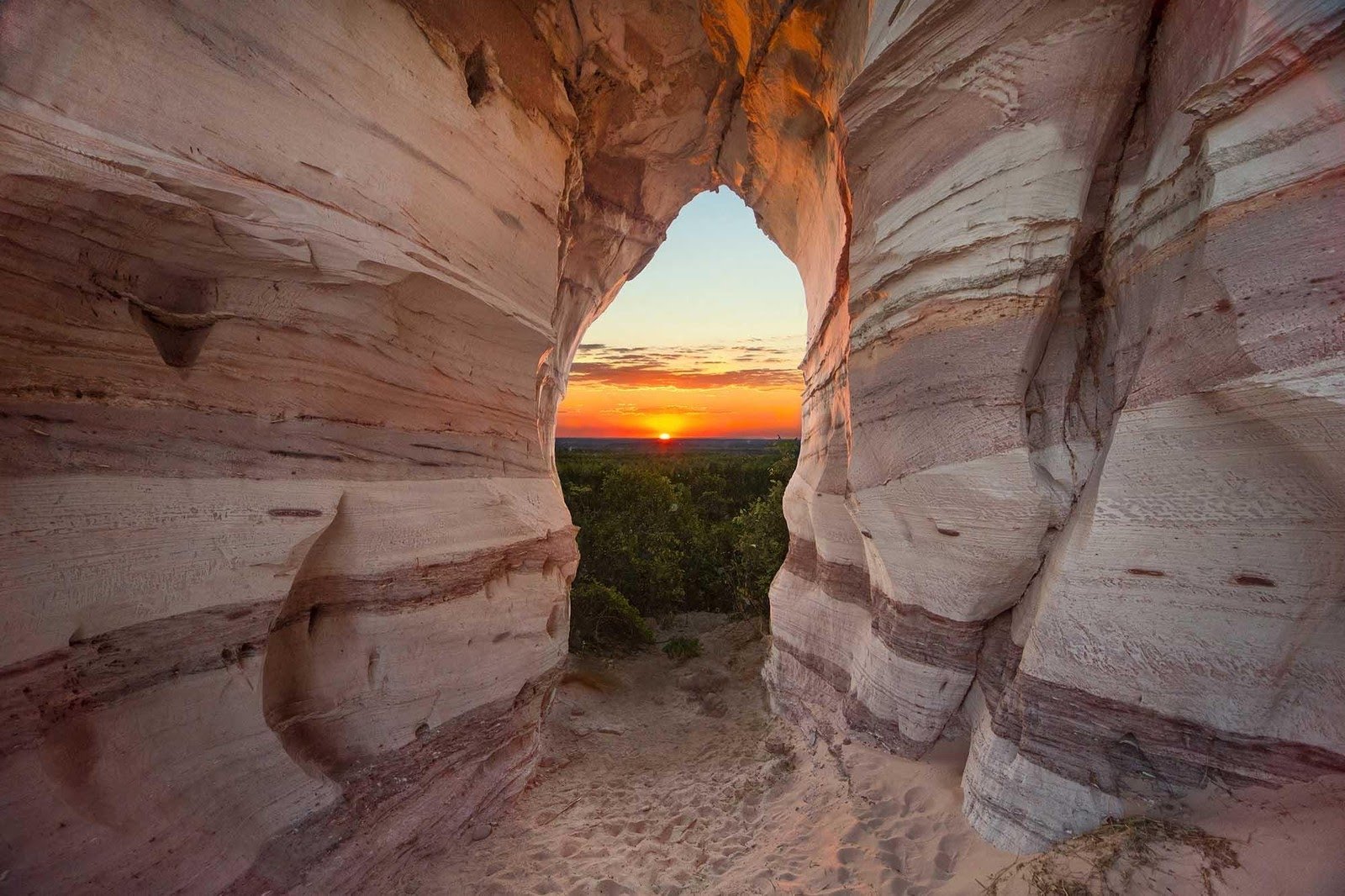 Pedra Furada als natuurlijk venster op een gloeiende ondergaande zon. Warm licht stroomt door een natuurlijke boog in gelaagd zandsteen tijdens een Jalapão-avontuur en omlijst een vurige zonsondergang boven een weids groen bladerdak