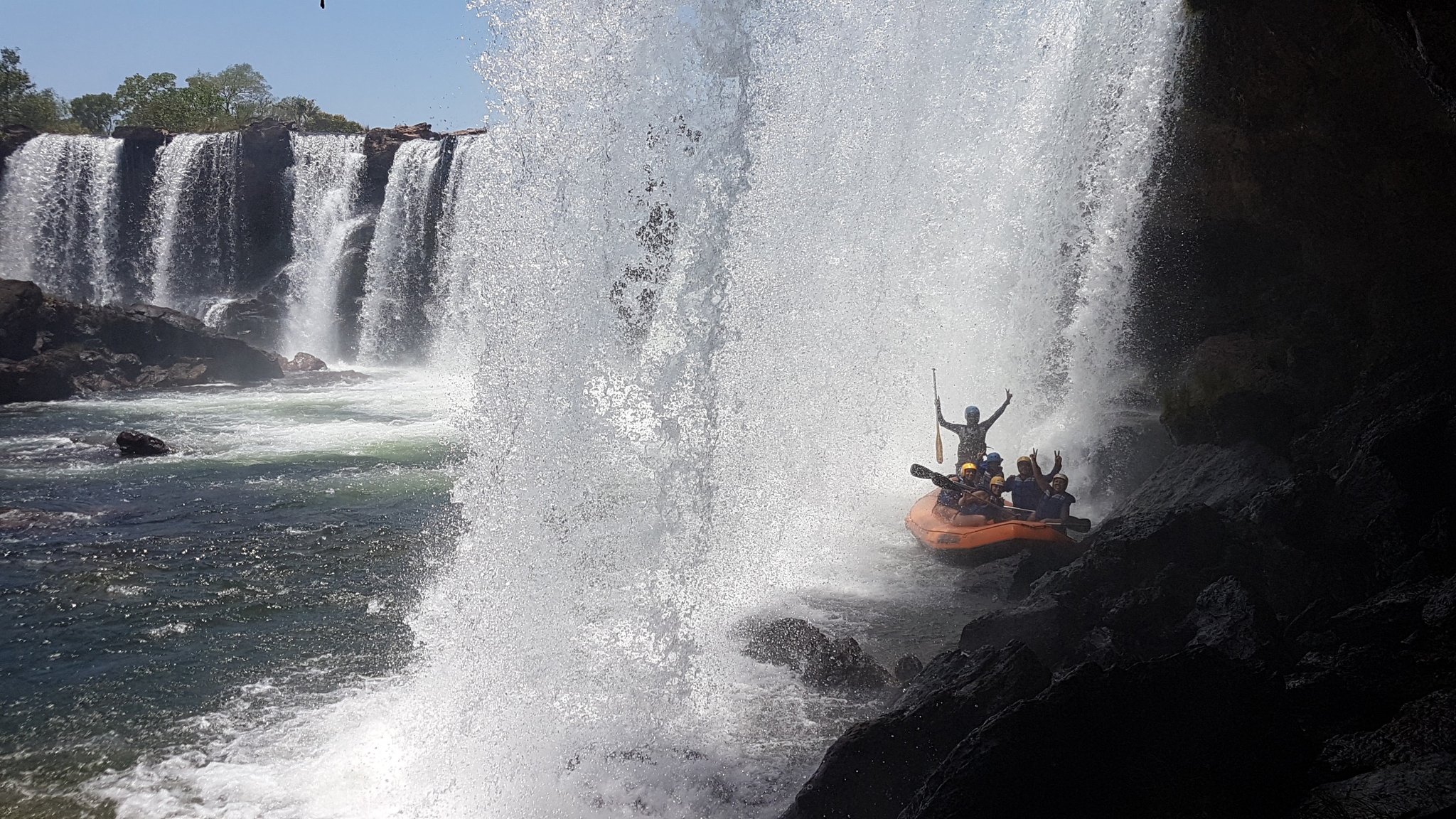 Doorweekt, juichend, armen omhoog — onder een waterval doorvaren is puur avontuur. Een groep behelmde raften in een oranje rubberboot viert feest onder een donderende waterval tijdens een Jalapão-avontuur, omringd door nevel en woest schuimend water