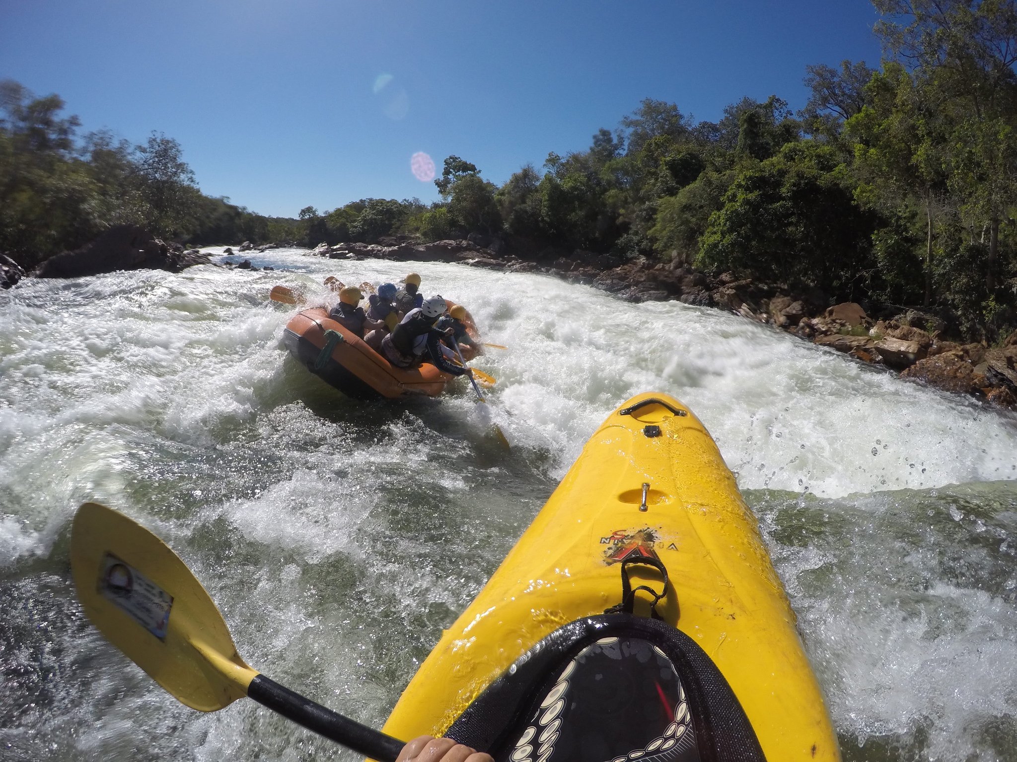 Paddling hard into the rapid — the Novo River delivers. POV from a yellow kayak on churning white-water rapids as a crew paddles an orange raft through a rocky gorge lined with green trees during Jalapão trekking