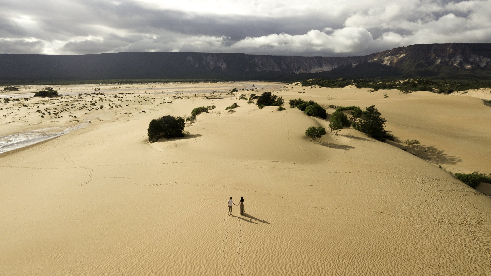 Two tiny figures, one vast dune field — footprints the only proof they were ever here. Photo: FG Trade Aerial view of two people holding hands on the sweeping golden sand dunes of Jalapão trekking country, with green shrubs, a distant river and dramatic sandstone cliffs under a cloudy sky