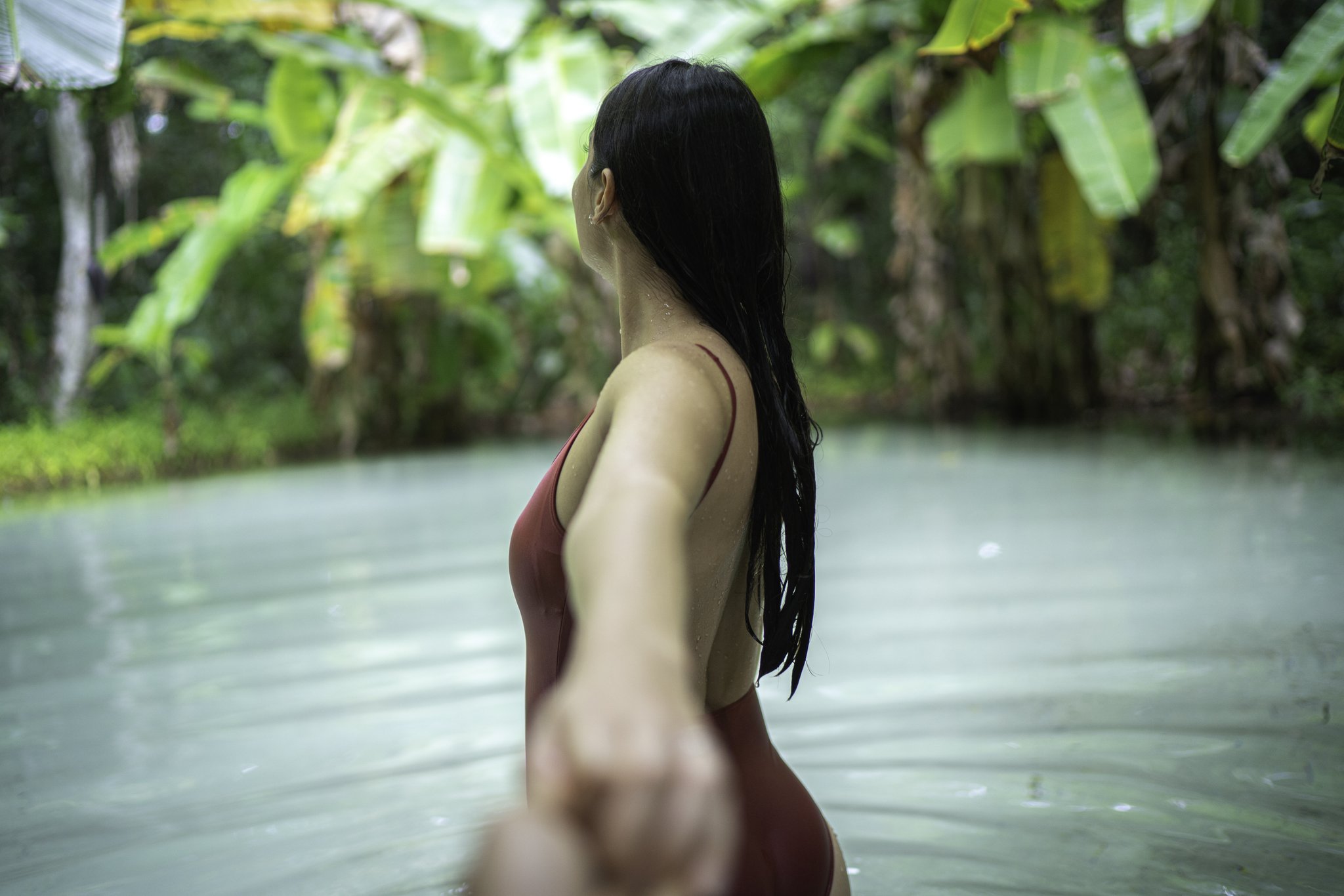 Cool, weightless water — a fervedouro moment worth every trail mile. Photo: FG Trade A woman in a red swimsuit wades through a pale turquoise natural spring pool surrounded by lush tropical vegetation, reaching back toward the camera during a Jalapão trekking experience