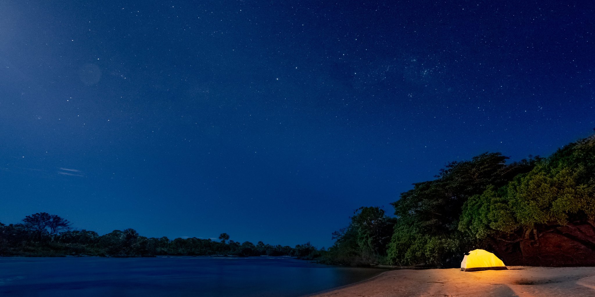 One glowing tent, a million stars — a night on the river beach. Photo: Alberto Andrich A single illuminated yellow tent pitched on a sandy river beach during a Jalapão trekking expedition, surrounded by dense tropical vegetation under a vast star-filled night sky