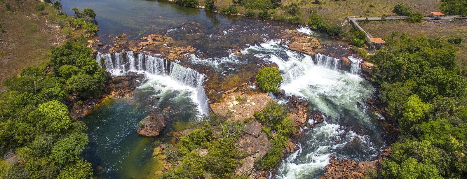 Scattered curtains of white water pour over ancient rock — Cachoeira da Velha from above. Aerial view of a wide horseshoe-shaped waterfall cascading over layered sandstone ledges into turquoise pools, surrounded by cerrado vegetation and a small viewing platform, on a Jalapão trekking route