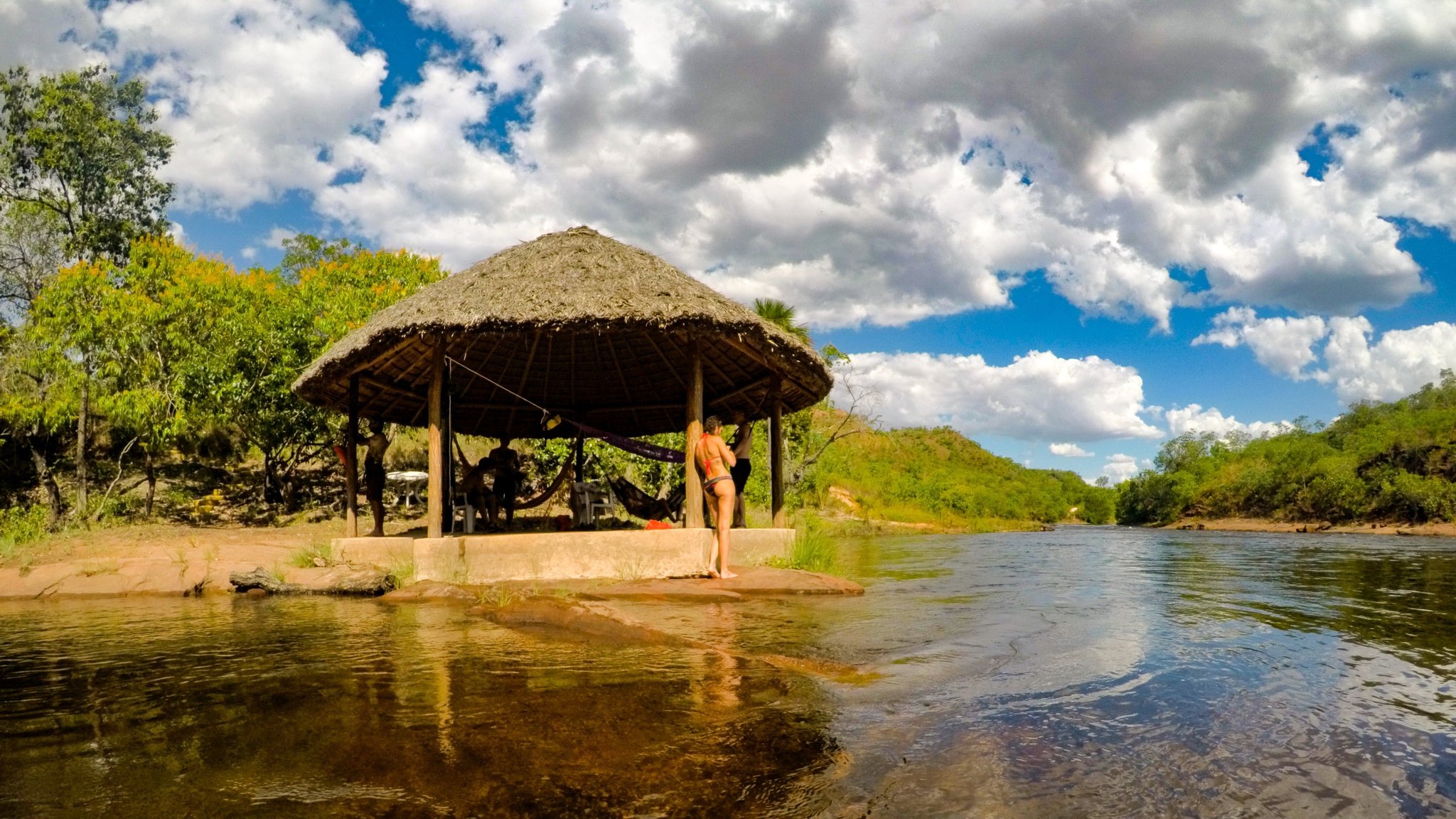 A shady palapa, hammocks, and cold river water — rest earned on the trail. Travelers relaxing under a round thatched-roof shelter on the banks of a clear river in Jalapão trekking country, with hammocks strung inside and green cerrado hills rising in the background