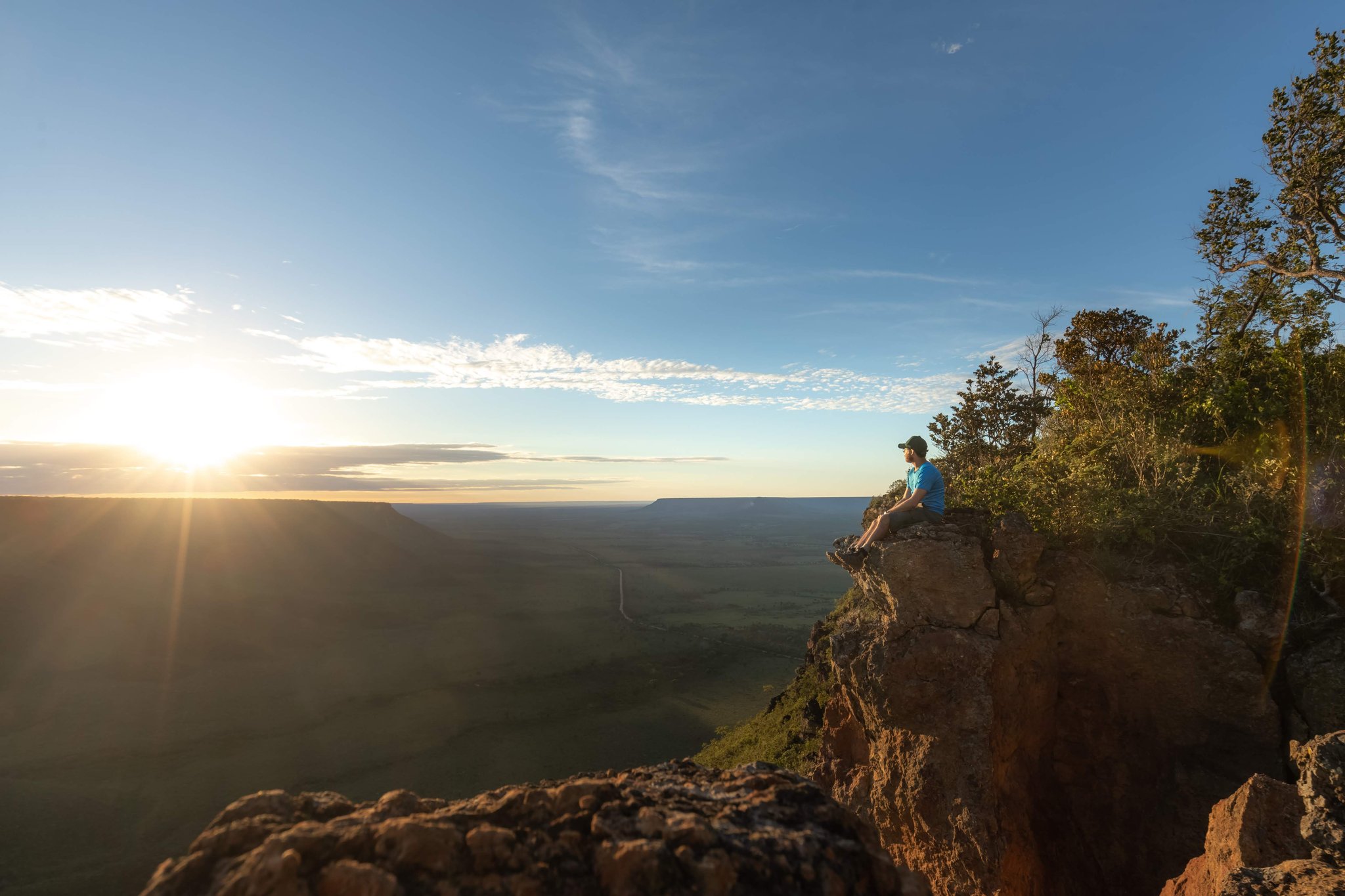 Sunrise from the plateau's edge — 200 metres above the world, and all of it yours. Photo: Alexandre Socci A lone traveler sits on the edge of a sandstone cliff during a Jalapão trekking expedition, gazing out over a vast green plateau bathed in golden sunrise light
