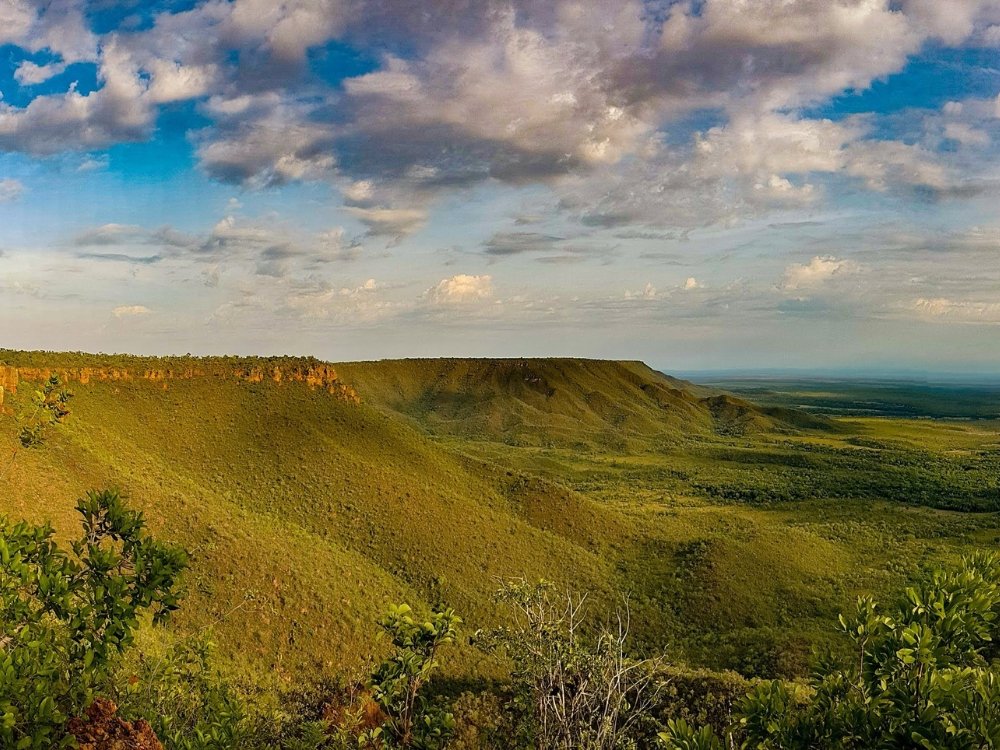 Summiting the plateau before the world wakes up — cerrado in every direction. Panoramic view from atop a sandstone plateau on a Jalapão trekking route, with green cerrado vegetation blanketing the slopes and a vast lowland plain stretching to the horizon under a dramatic cloudy sky