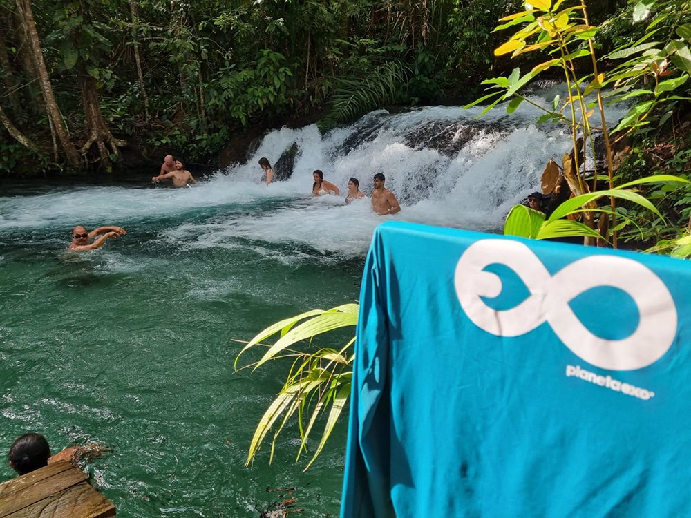 Rushing white water, cool turquoise pools — earned with every step. A group of travellers swimming and wading at the base of a cascading waterfall surrounded by dense tropical forest on a Jalapão trekking experience, with a PlanetaEXO branded towel in the foreground
