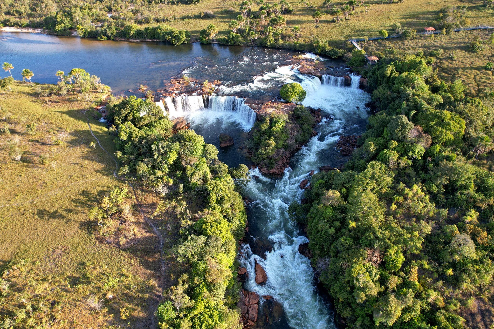 Três quedas, uma corrida furiosa pelas rochas do cerrado. Photo: Wirestock Vista aérea de uma cachoeira escalonada no Jalapão, com corredeiras brancas cortando canais rochosos, rodeada por cerrado e um lago calmo a montante