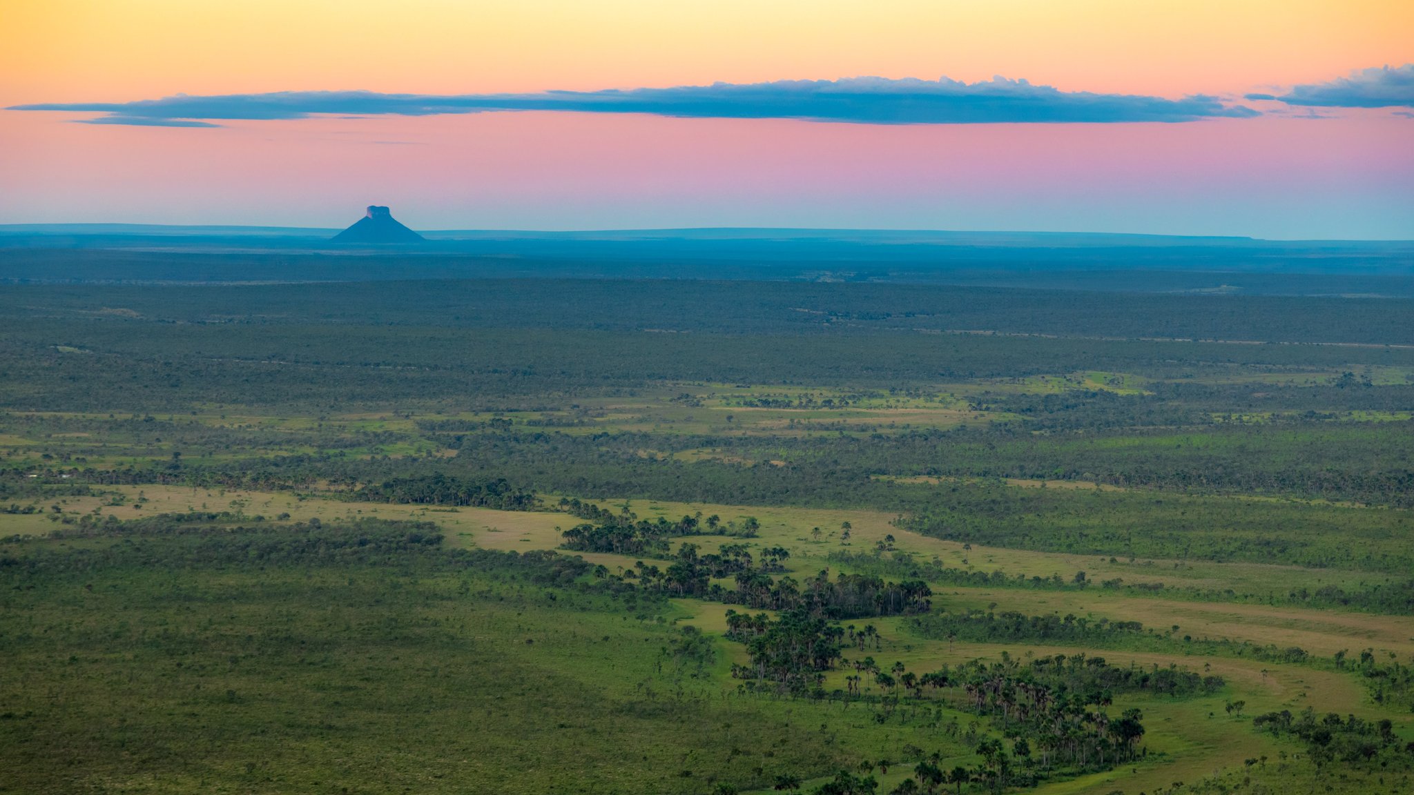 Savana sem fim, um morro solitário, céu que não sossega. Photo: Alberto Andrich Vasta savana do Cerrado estendendo-se até o horizonte em uma viagem ao Jalapão, com um morro de topo plano em silhueta sob um céu crepuscular rosa e âmbar
