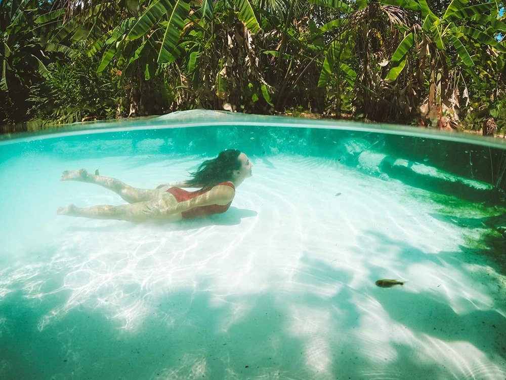 Flutuando no azul-turquesa sem tocar o fundo — isso só existe no Jalapão. Uma mulher de biquíni vermelho desliza sob a superfície turquesa cristalina de um fervedouro em uma viagem ao Jalapão, com fundo de areia branca e vegetação tropical exuberante visível acima da linha d'água
