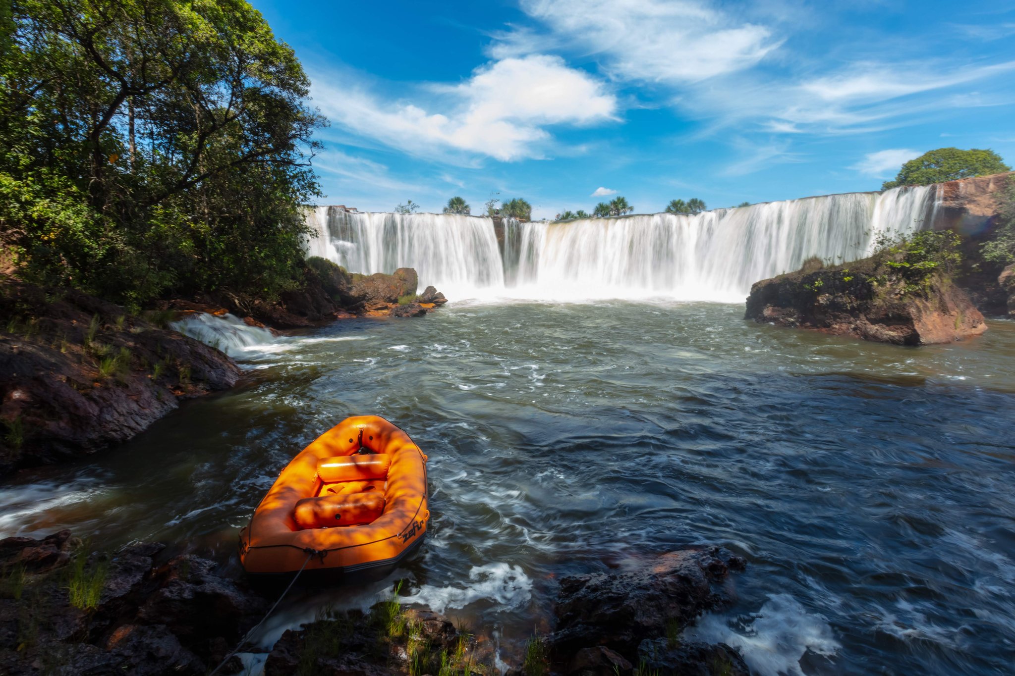Bote parado, cachoeira trovejando — o Rio Soninho reserva emoções. Photo: Alberto Andrich Um bote inflável laranja ancorado em margem rochosa diante de uma ampla cachoeira em viagem ao Jalapão, cercado por vegetação de cerrado sob céu azul