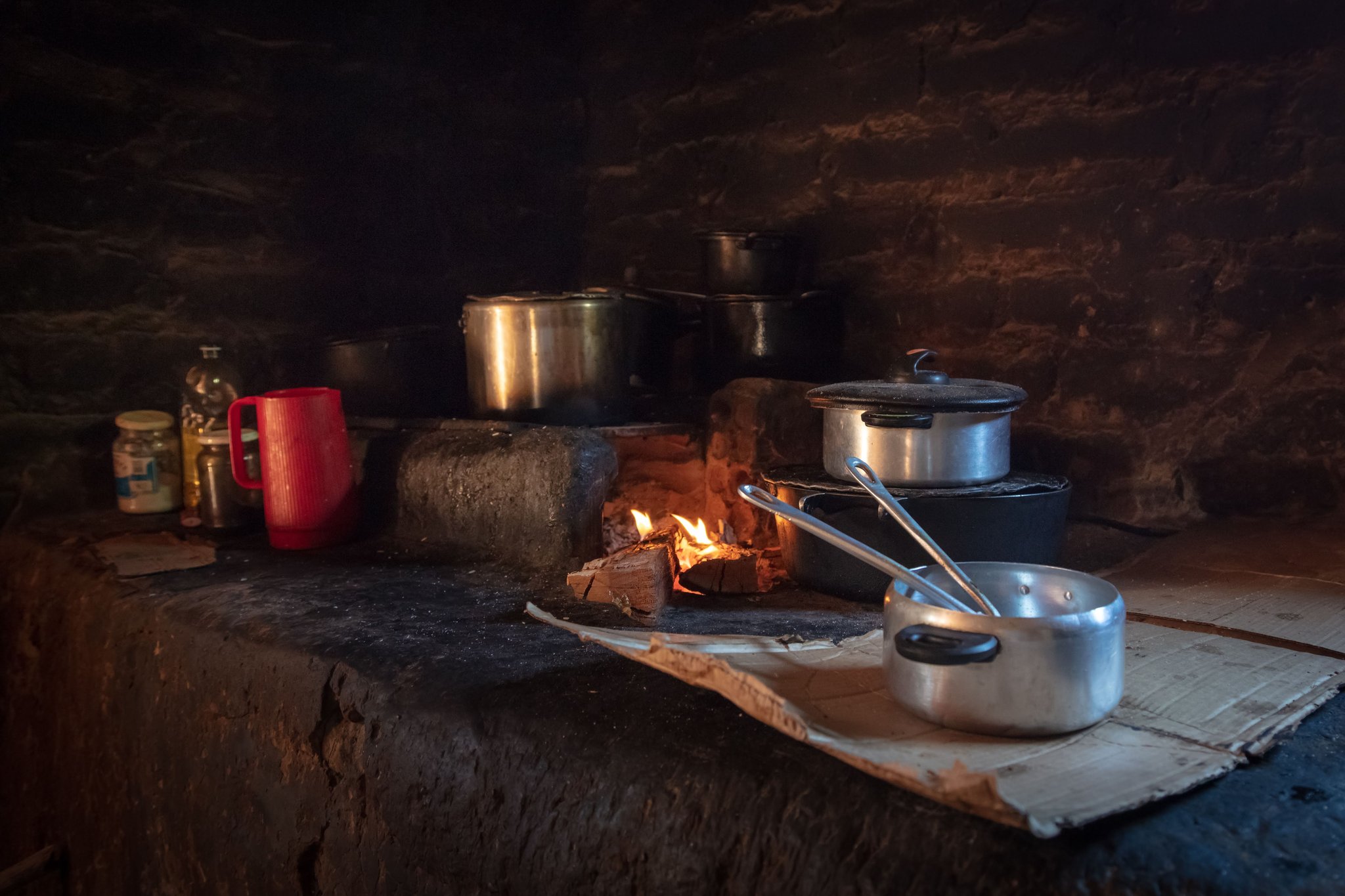 Várias panelas e frigideiras de alumínio fervendo sobre fogo a lenha em um fogão de pedra rústico, com uma caneca vermelha e potes de condimentos em uma cozinha tradicional pouco iluminada