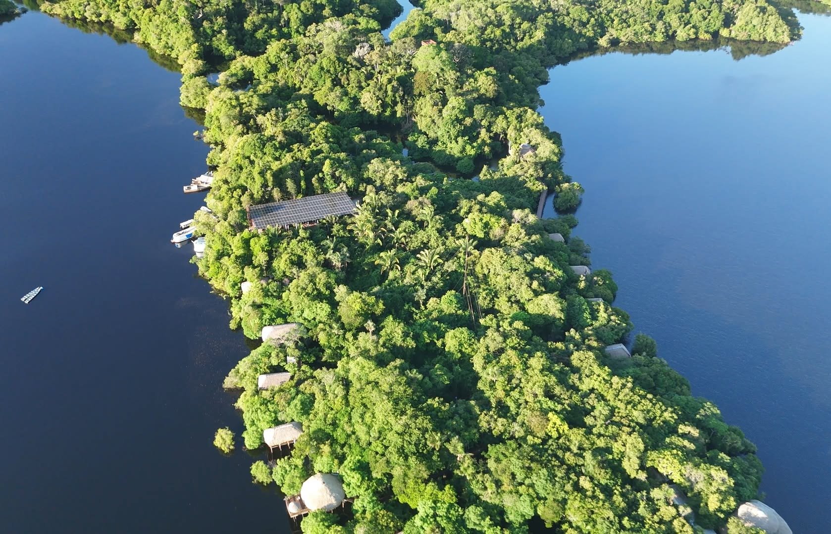 Luftaufnahme der Juma Amazon Jungle Lodge auf einer dicht bewaldeten Flussinsel, umgeben von dunklem Amazonaswasser, mit Strohdach-Bungalows und Solarpanelen im Blätterdach