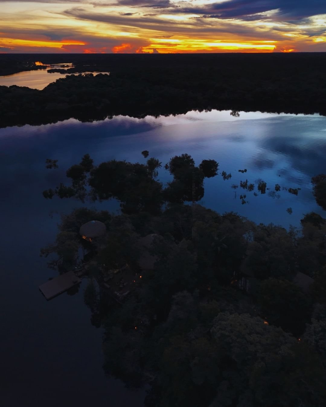 Drohnenaufnahme der Juma Amazon Jungle Lodge bei Einbruch der Dunkelheit: warme Lichter zwischen überschwemmten Waldinselchen und ein spektakulärer orange-goldener Sonnenuntergang, der sich im dunklen Fluss spiegelt