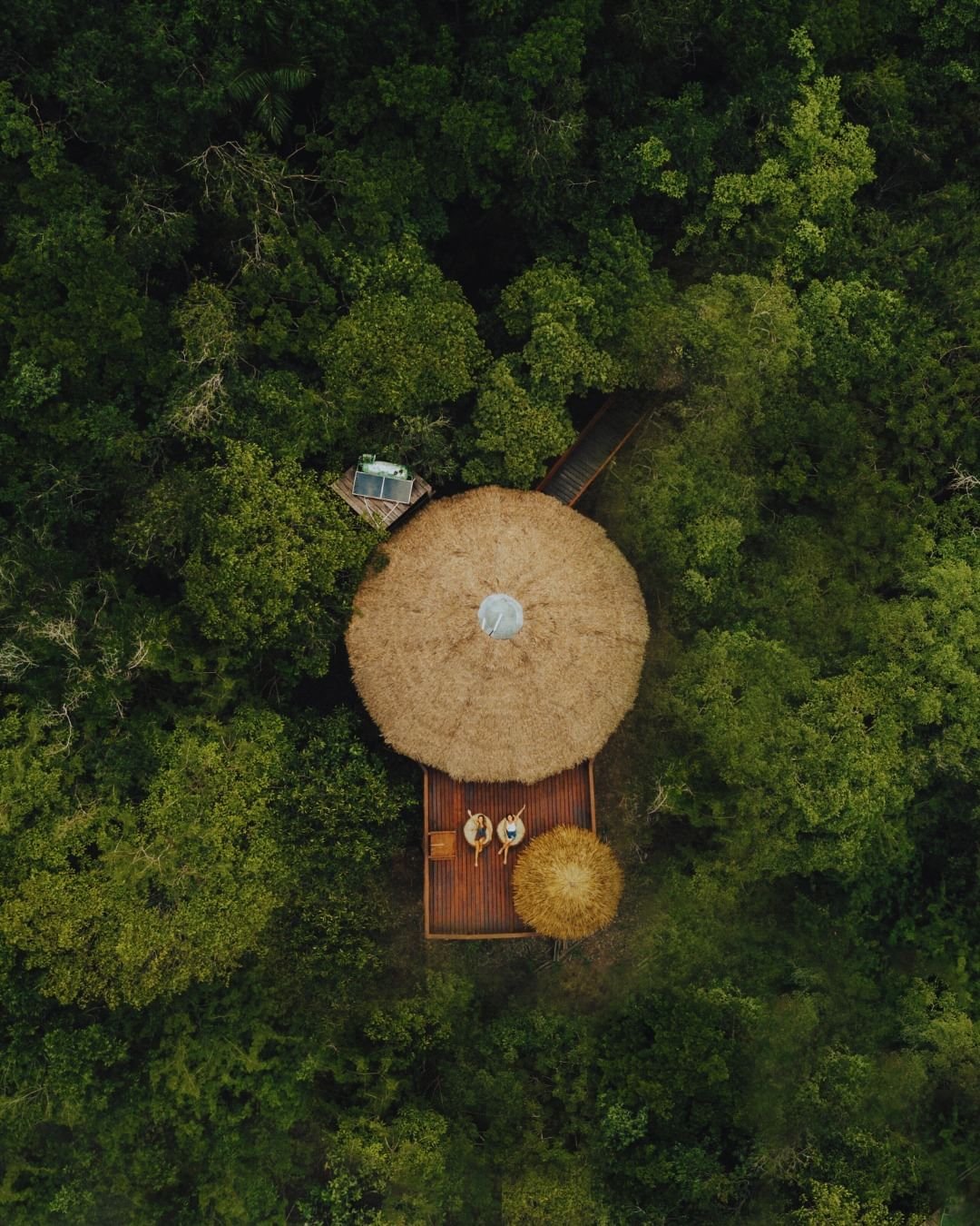 Luftaufnahme eines runden Bungalows mit Strohdach in der Juma Amazon Jungle Lodge, umgeben von dichtem Regenwalddach, mit zwei Gästen auf einer Holzterrasse