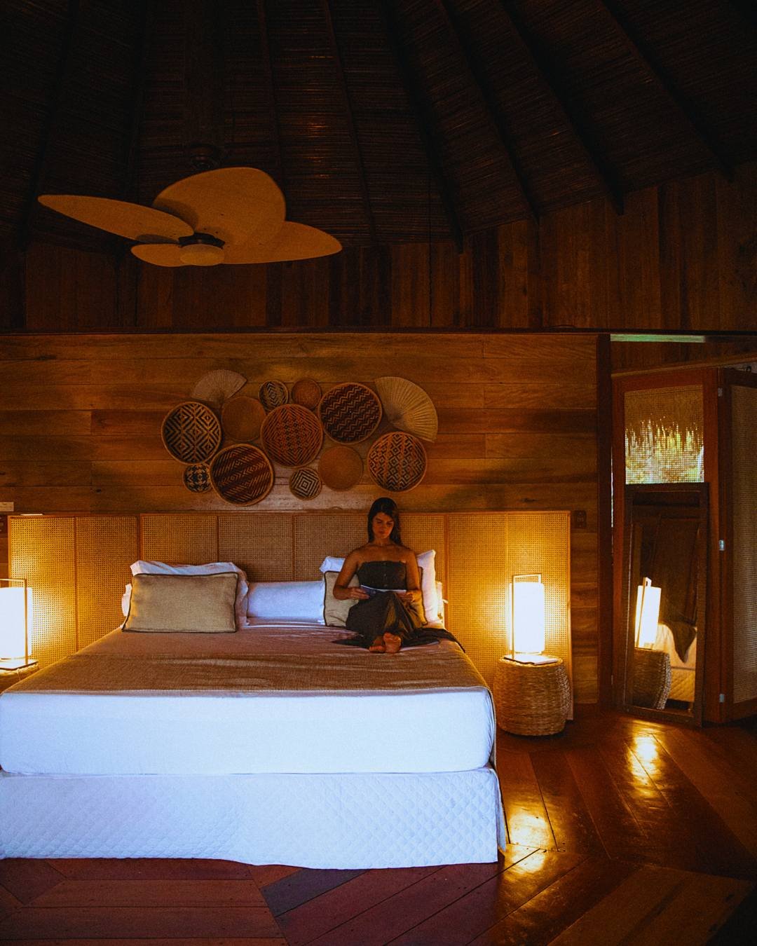 Eine Frau liest auf einem Kingsize-Bett im Bungalow der Juma Amazon Jungle Lodge – warmes Lampenlicht, ein geflochtenes Rattankopfteil, dekorative Körbe an Holzwänden und eine Reetdecke mit Holzventilator