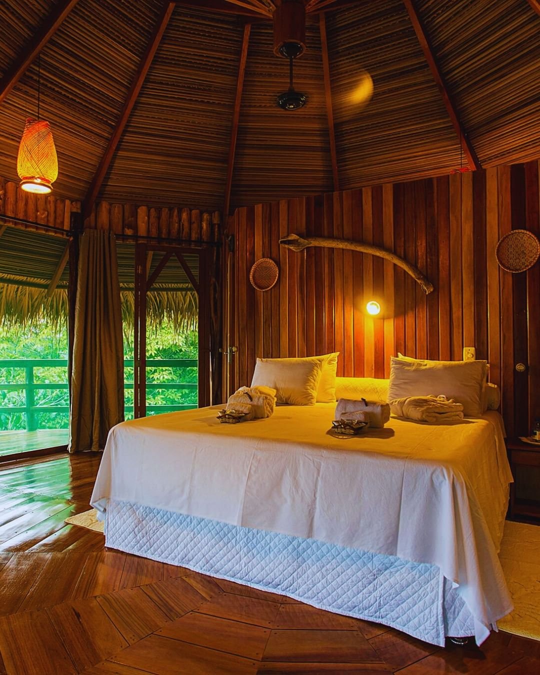 Warmes Bungalow-Interieur der Juma Amazon Jungle Lodge mit Doppelbett in weißer Bettwäsche, kegelförmigem Strohdach, poliertem Hartholzboden und einem offenen Balkon mit Blick auf üppige Regenwaldvegetation