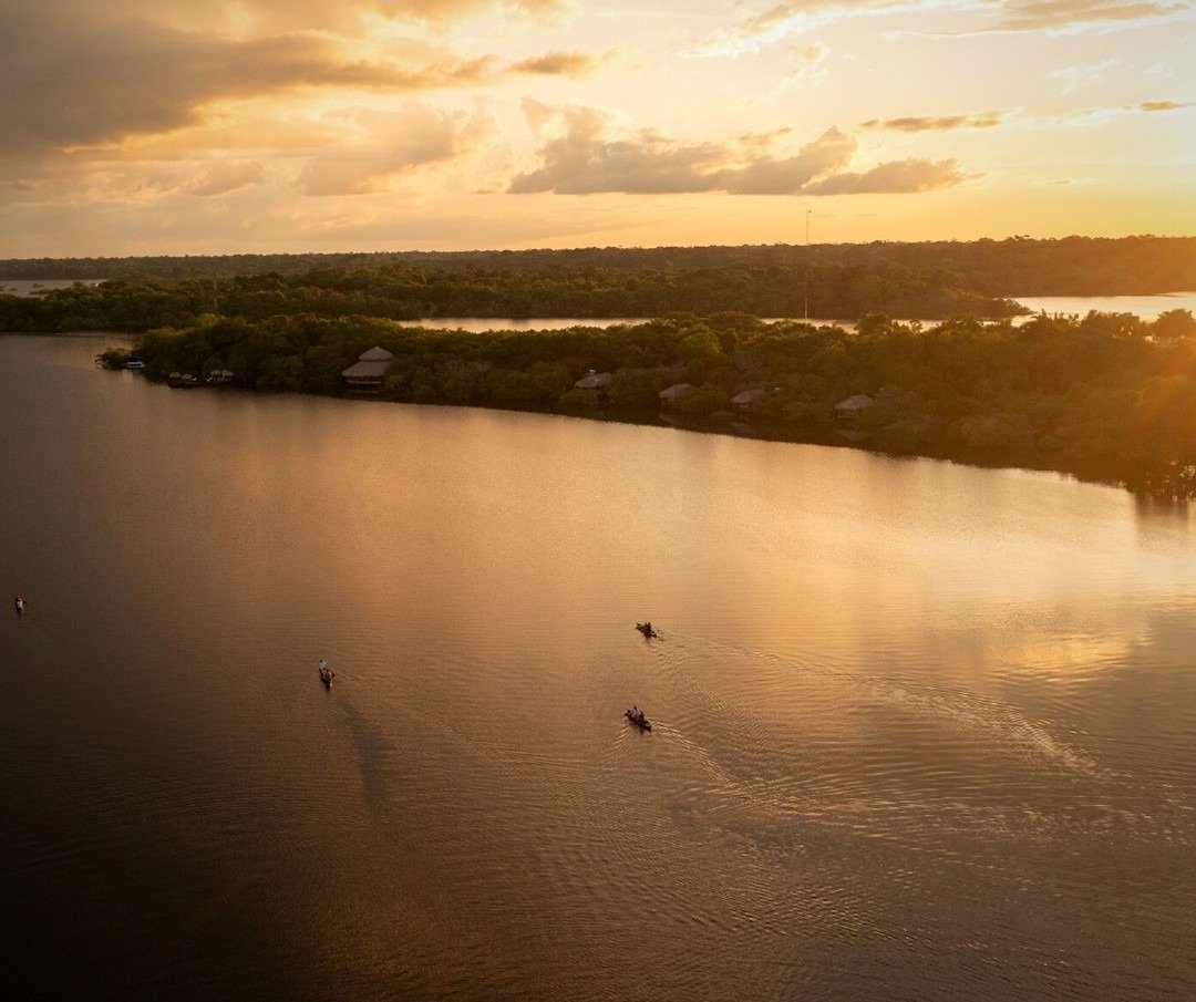 Luftaufnahme kleiner Kanus auf einem breiten Fluss im goldenen Sonnenuntergangslicht, mit den Strohdach-Bungalows der Juma Amazon Jungle Lodge im dichten Regenwald am gegenüberliegenden Ufer