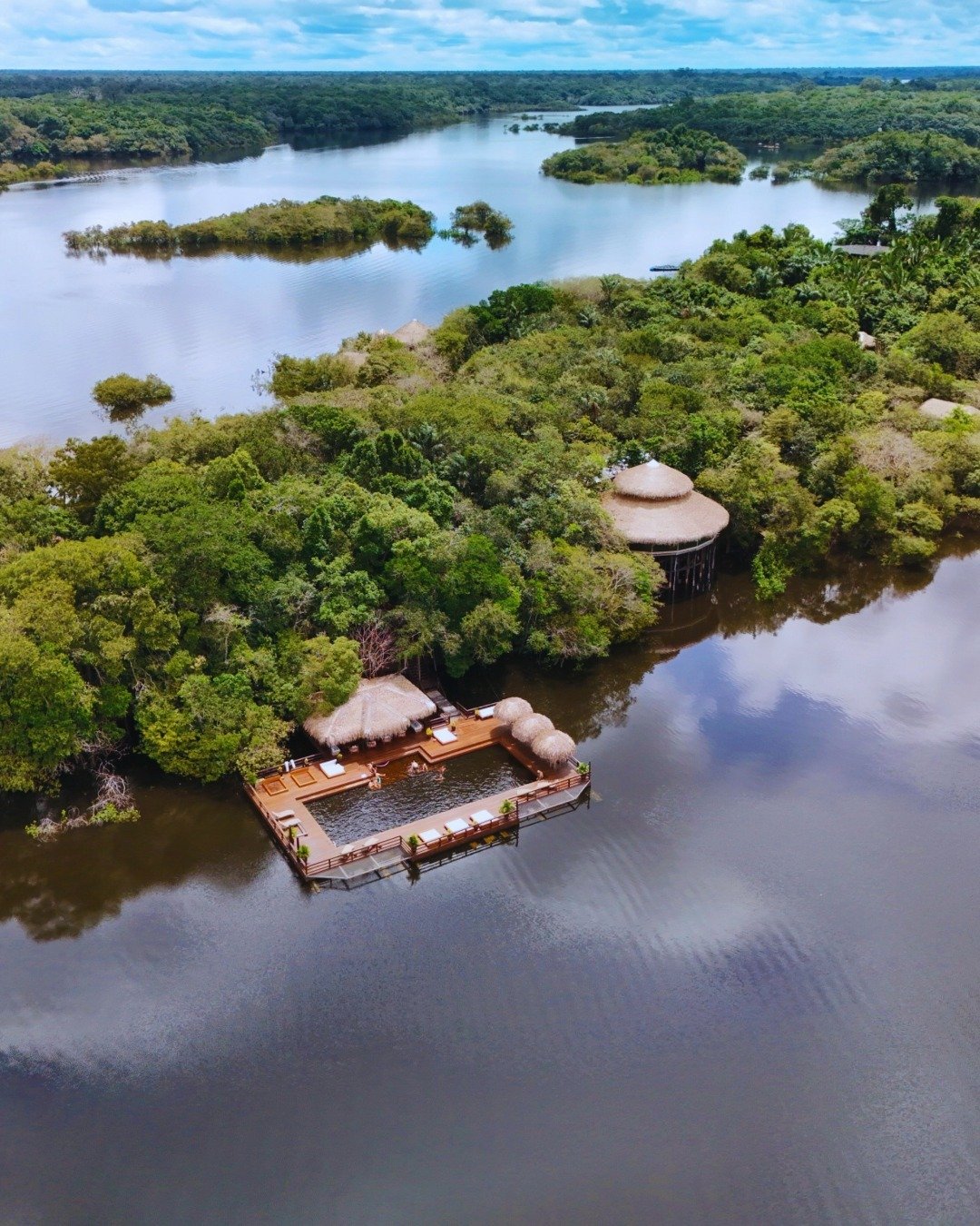 Luftaufnahme der Juma Amazon Jungle Lodge mit einem schwimmenden Holzdeck, strohgedeckten Pavillons und einem Flusspool inmitten überschwemmten Regenwaldes