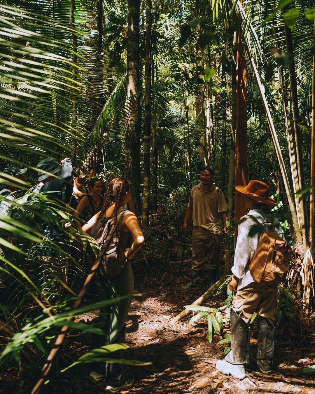 Eine kleine Gruppe lächelnder Reisender folgt einem einheimischen Führer auf einem schmalen Pfad durch dichten tropischen Regenwald der Juma Amazon Jungle Lodge, umgeben von Palmwedeln und hohen Bäumen