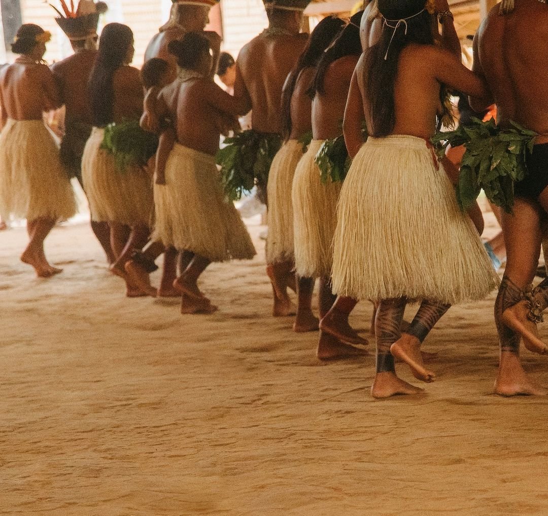 Eine Reihe indigener Tänzerinnen und Tänzer in geflochtenen Strohröcken und Blattschmuck bewegt sich barfuß über einen Sandboden bei einer traditionellen Zeremonie während des Gemeinschaftsbesuchs in der Juma Amazon Jungle Lodge