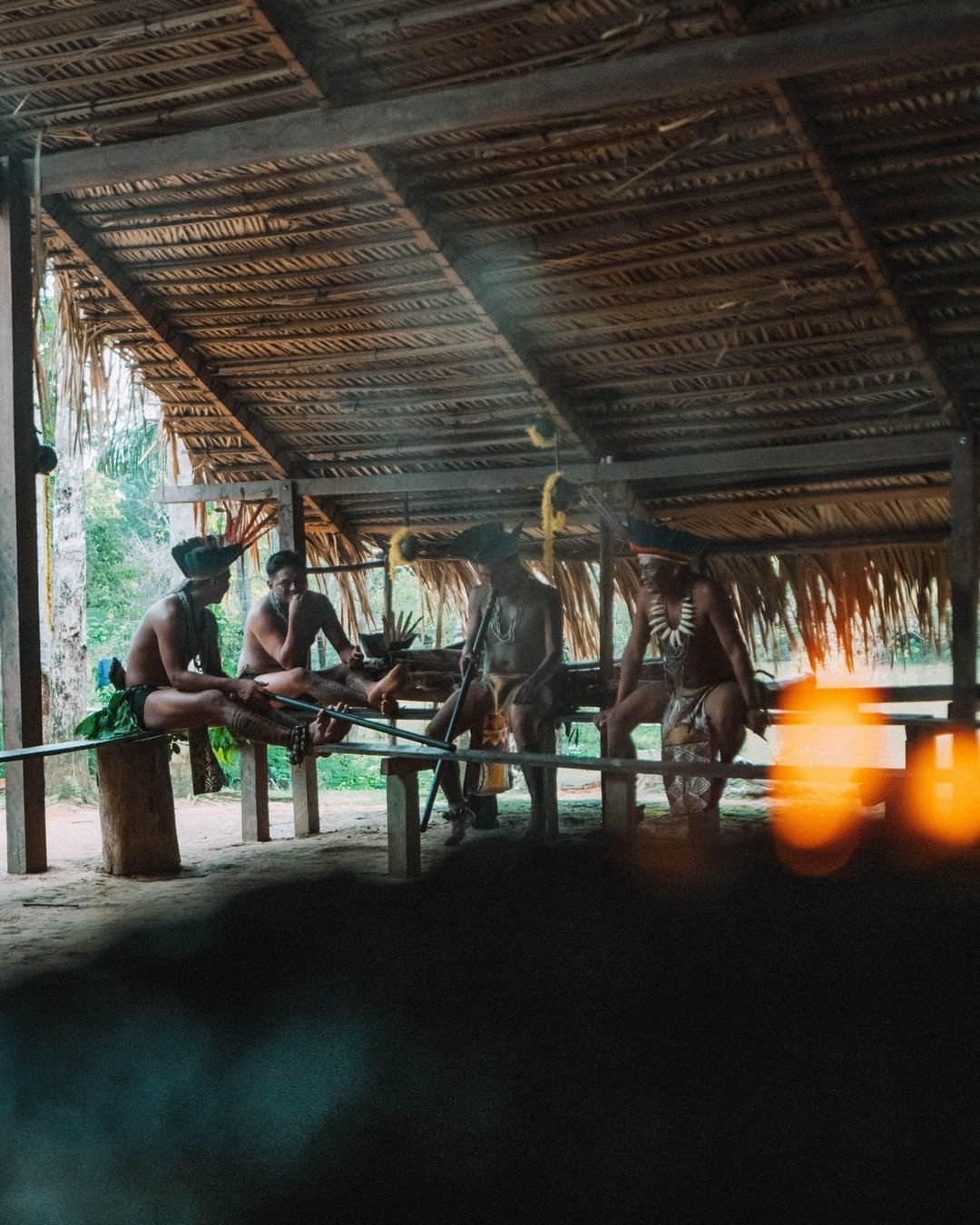 Mitglieder einer indigenen Gemeinschaft in traditioneller Körperbemalung, Kopfschmuck und Perlenketten versammeln sich unter einem strohgedeckten Freiluftgebäude der Juma Amazon Jungle Lodge, lange Blasrohre lehnen auf Holzbänken