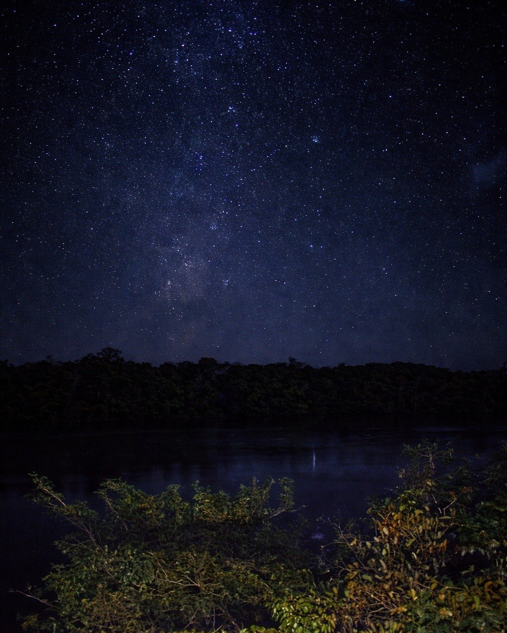 Countless stars, total silence — the Amazon night sky from the water's edge. The Milky Way stretches across a star-filled sky above a dark jungle river at Juma Amazon Jungle Lodge, with the galactic core faintly reflected in the still water below