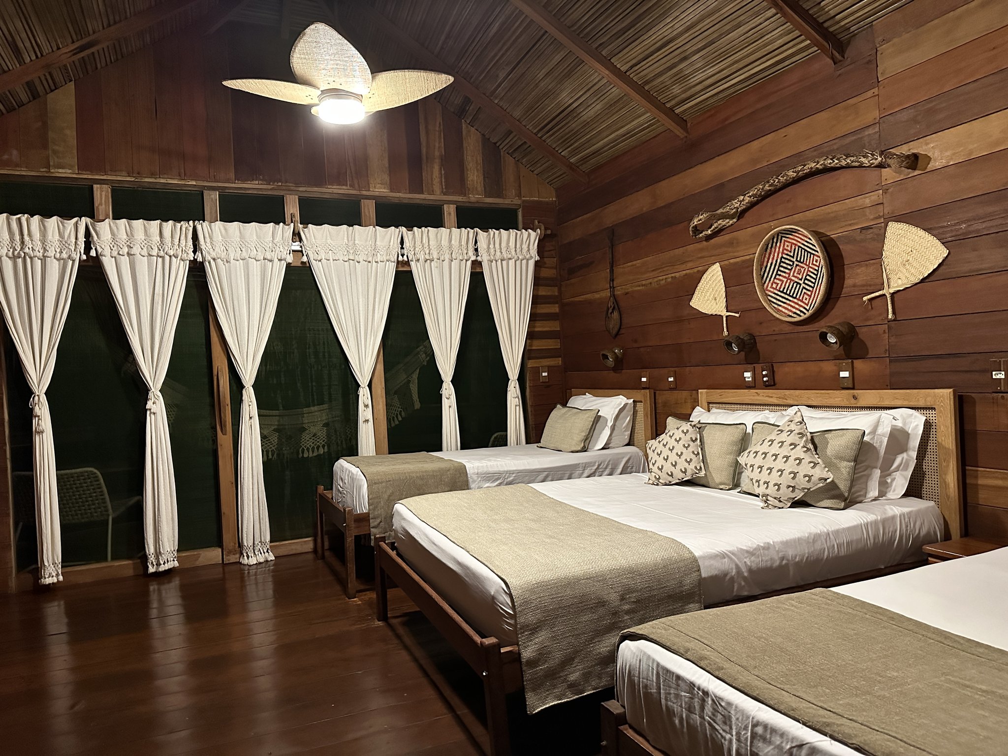 Geräumiges Bungalow der Juma Amazon Jungle Lodge bei Nacht mit drei Holzbetten in weißer Bettwäsche, einem geflochtenen Deckenventilator und einheimischen Korbwaren und Paddeln an den Holzwänden