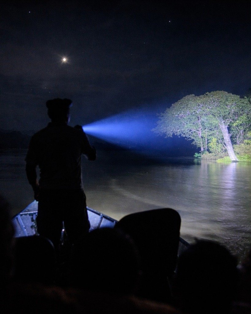 Silhouette eines Guides, der auf dem Bug eines kleinen Bootes steht und bei einer Nachttour der Juma Amazon Jungle Lodge einen hellen Scheinwerfer über einen dunklen Fluss auf einen großen Baum richtet, während ein Halbmond am Sternenhimmel leuchtet