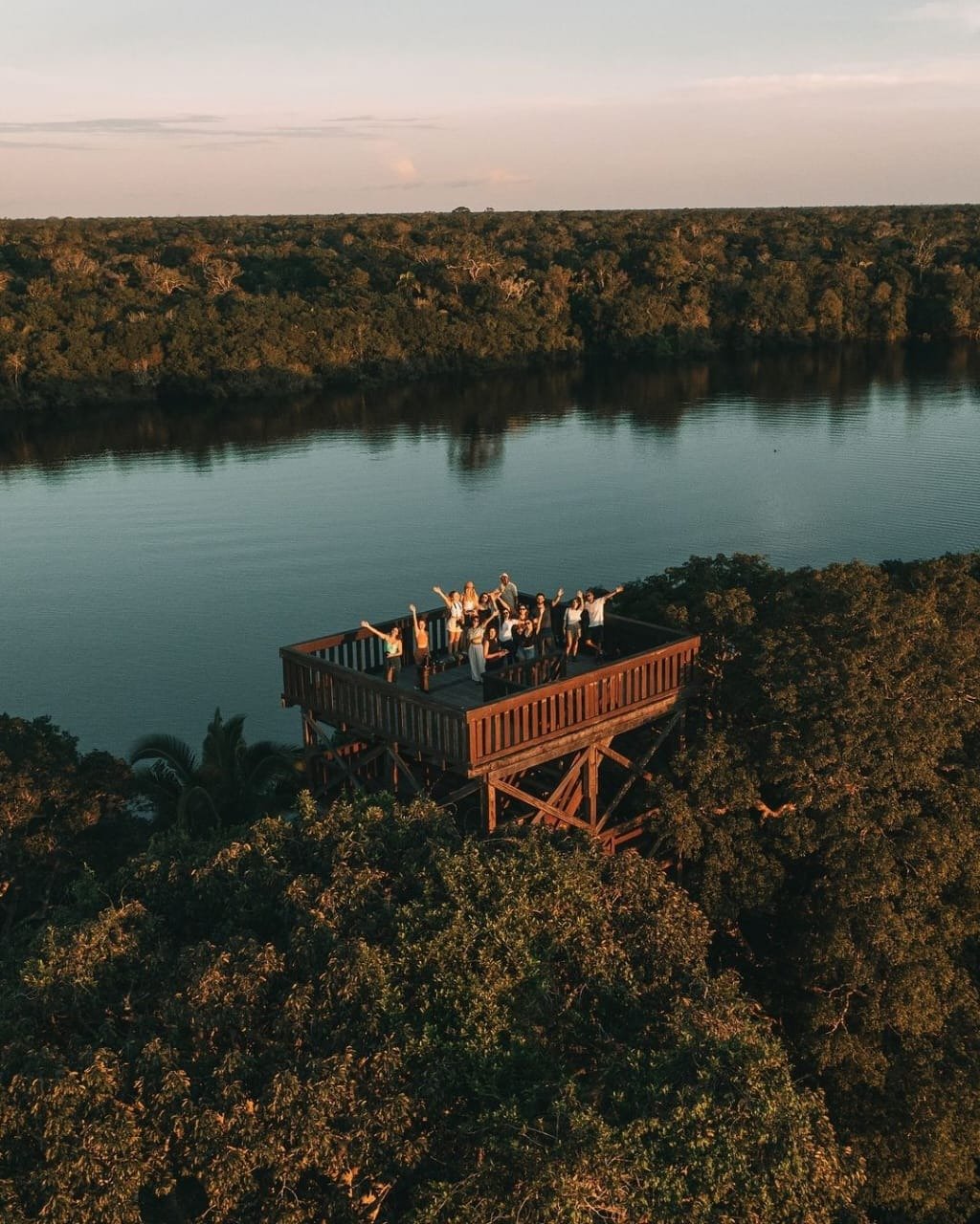 Eine Reisegruppe winkt begeistert von einem hohen hölzernen Aussichtsturm der Juma Amazon Jungle Lodge, umgeben von dichtem Regenwalddach und einem ruhigen Fluss im Sonnenuntergang