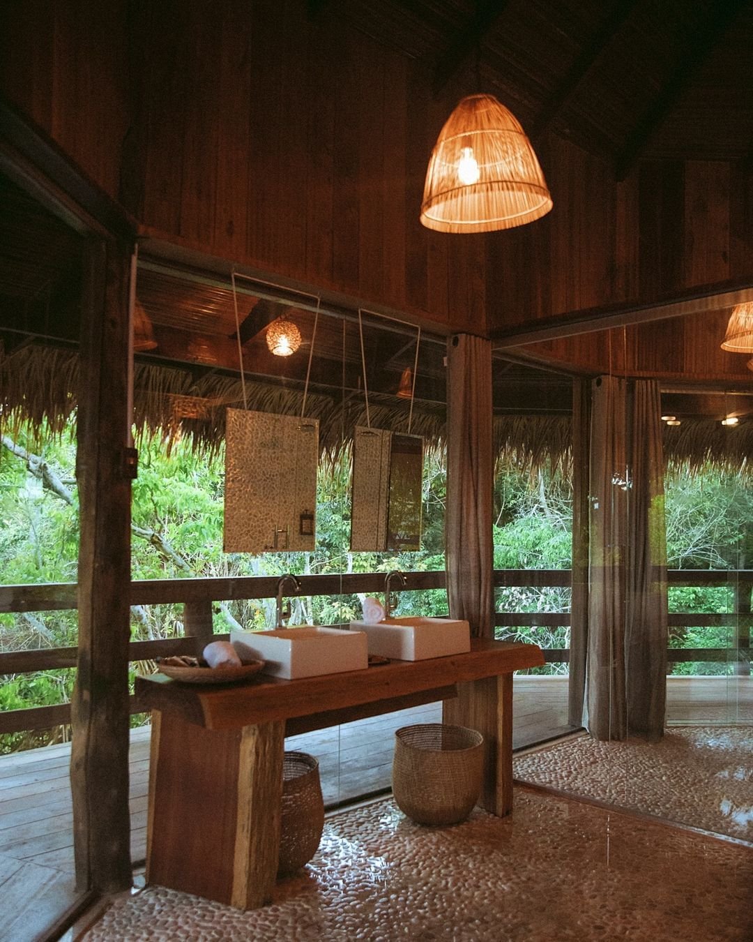 Offenes Badezimmer in der Juma Amazon Jungle Lodge mit zwei Aufsatzwaschbecken auf einem Massivholz-Waschtisch, geflochtenem Hängelampenschirm, Kieselsteinboden und raumhohen Glaswänden mit Blick in den Regenwald