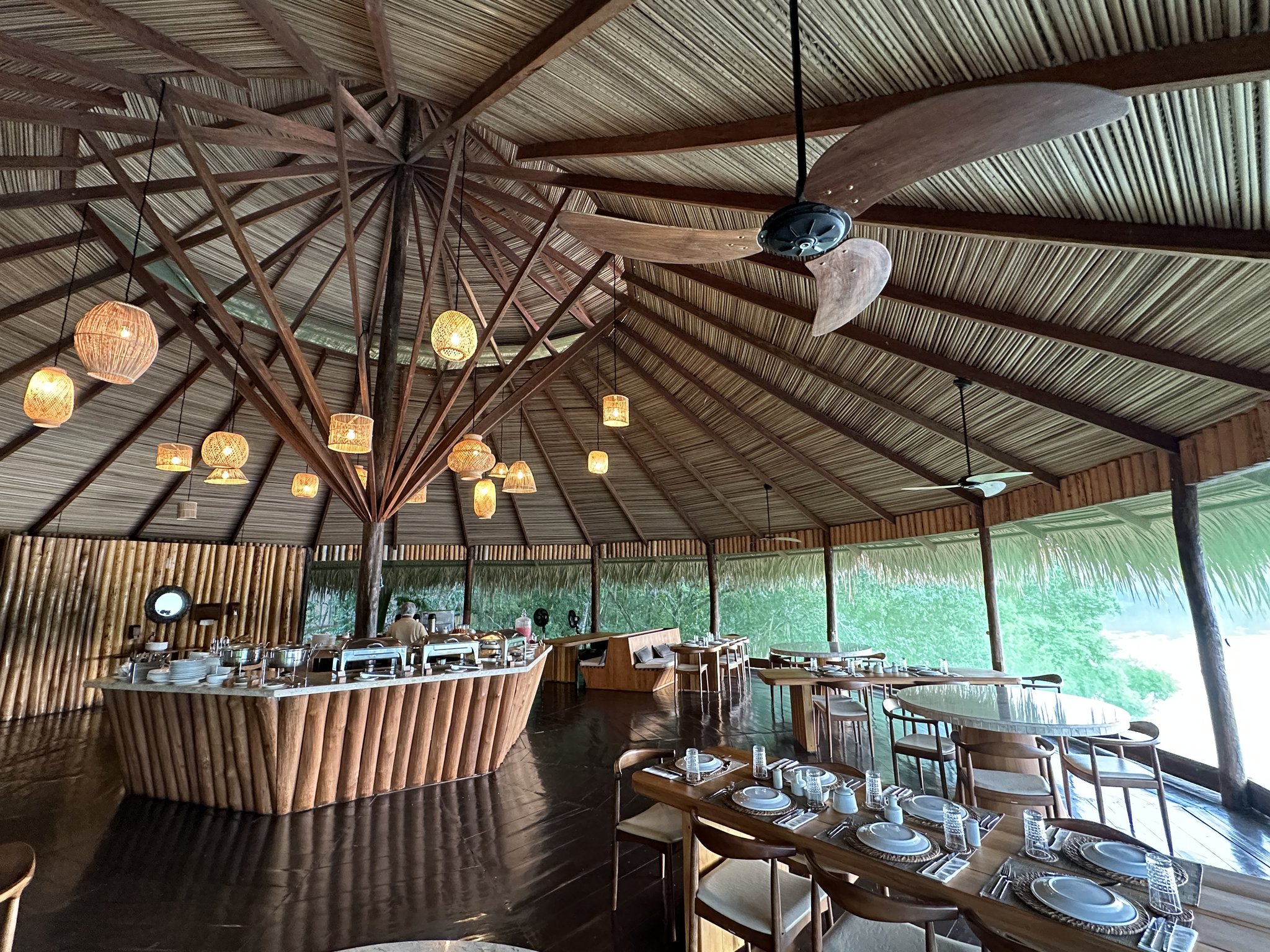 Das Open-Air-Restaurant des Juma Amazon Jungle Lodge mit einem hohen Strohdach aus radialen Holzbalken, geflochtenen Rattanleuchten, einer bambusverkleideten Buffettheke und gedeckten Holztischen, umgeben von üppigem Regenwald