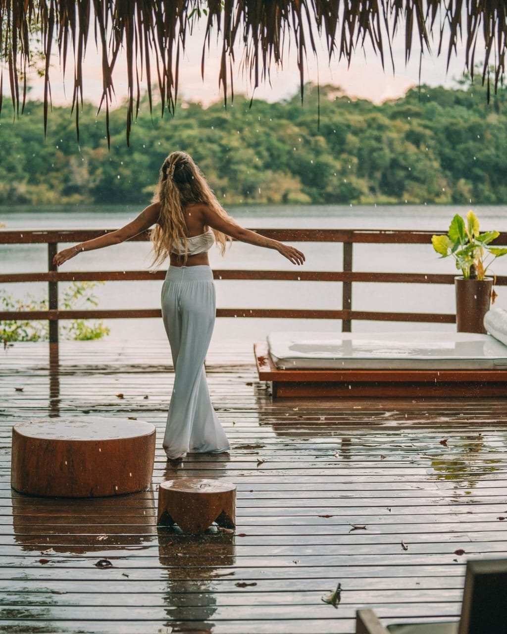 Eine Frau in Weiß steht mit ausgebreiteten Armen auf einer regenfeuchten Terrasse der Juma Amazon Jungle Lodge und blickt auf einen ruhigen Fluss mit dichtem Regenwaldufer