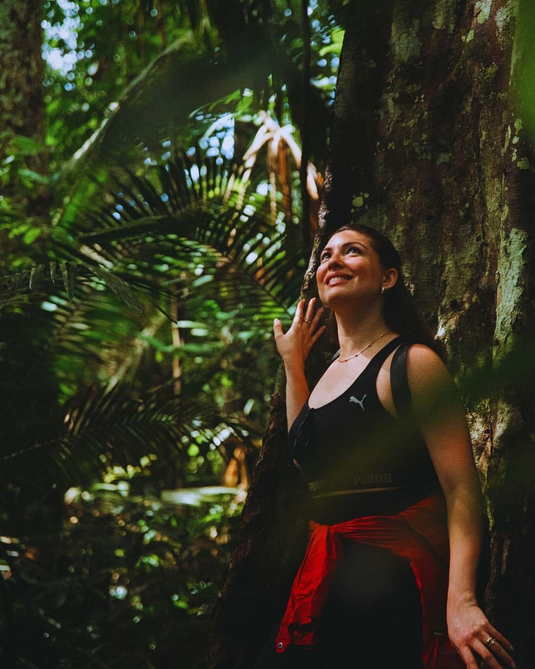 Leaning into the Amazon — every trunk tells a century of stories. A smiling woman leans against a massive rainforest tree trunk on a Juma Amazon Jungle Lodge guided hike, surrounded by lush ferns and towering palms