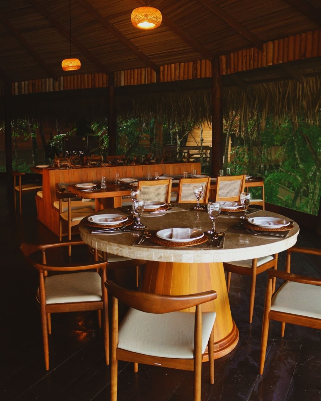 Gedeckter ovaler Esstisch mit geflochtenen Untersetzern, Weingläsern und weißen Tellern im offenen Restaurant der Juma Amazon Jungle Lodge, umgeben von Bambuswänden, einem Strohdach und üppigem Regenwald