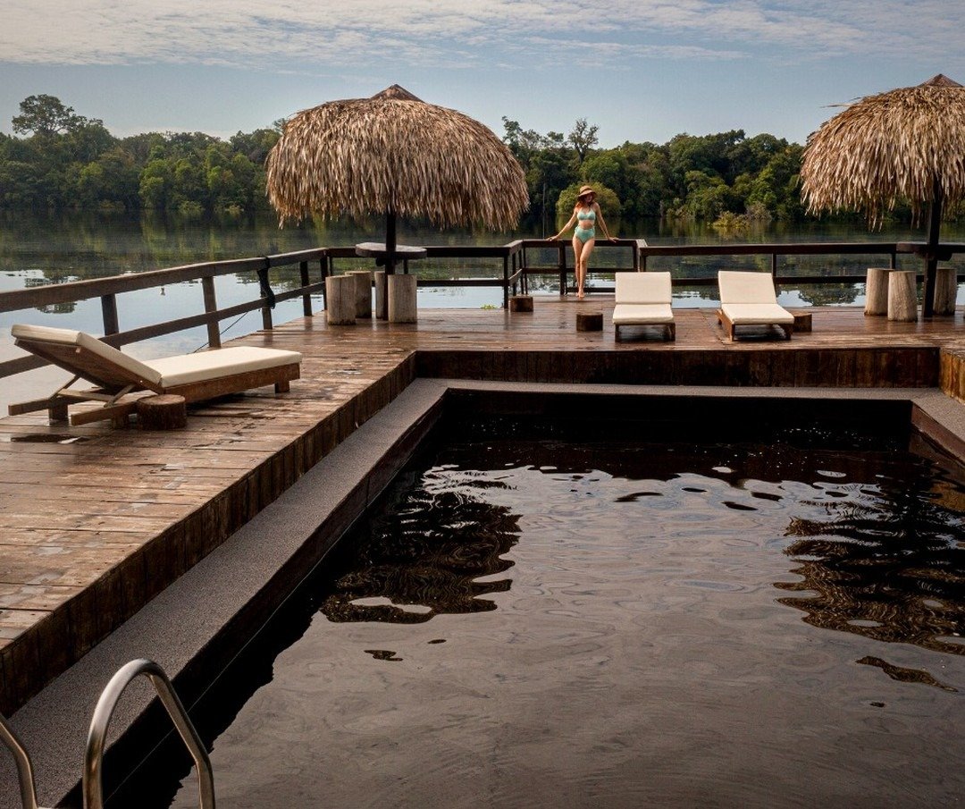 Ein Fluss-Pool mit dunklem Wasser auf einem Holzdeck der Juma Amazon Jungle Lodge, mit Strohdach-Sonnenschirmen, Liegestühlen und einer Gästin mit Blick auf den ruhigen Dschungelfluss