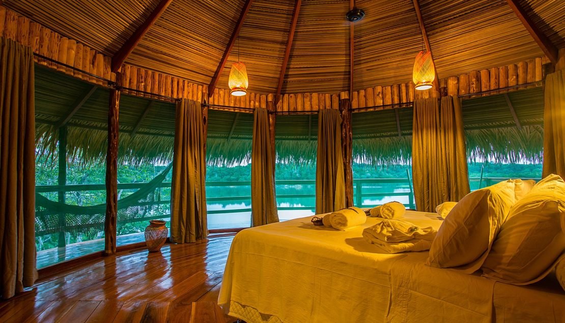 Inneres eines Bungalows im Juma Amazon Jungle Lodge mit Doppelbett, geflochtenen Hängelampen, Reetdach und offenen Fenstern mit Blick auf einen ruhigen Dschungelfluss