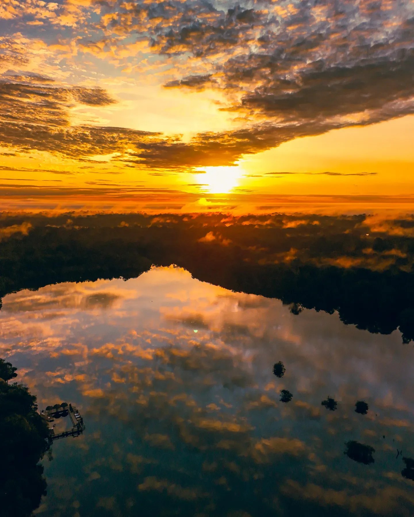 Luftaufnahme eines leuchtend goldenen Sonnenaufgangs über einem Regenwaldsee bei der Juma Amazon Jungle Lodge, mit dramatischen orangefarbenen Wolken, die sich im spiegelglatten Wasser spiegeln