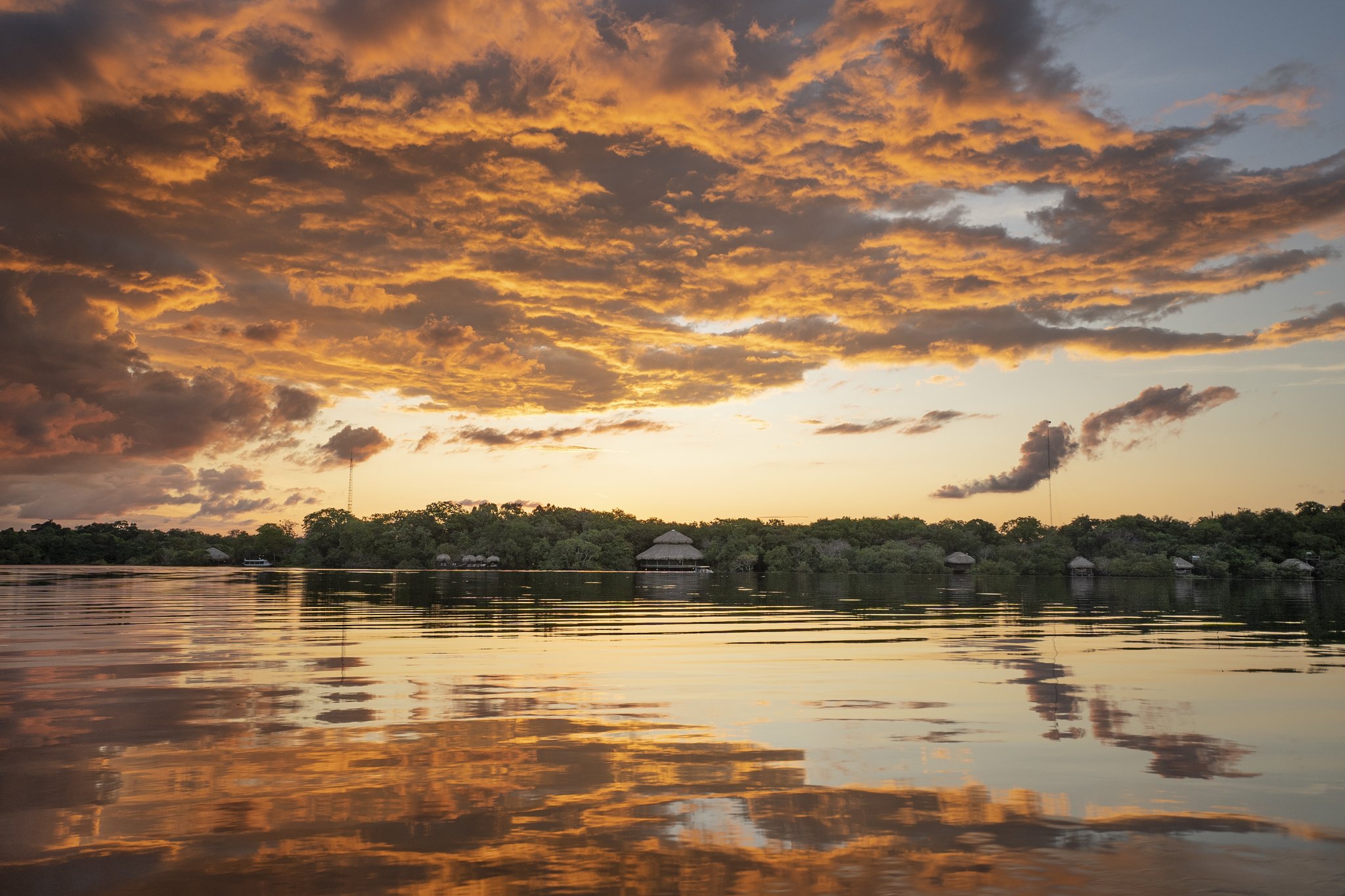 A fiery sunset sky reflected on a still river in front of the Juma Amazon Jungle Lodge, with thatched-roof bungalows nestled among dense rainforest trees along the far bank
