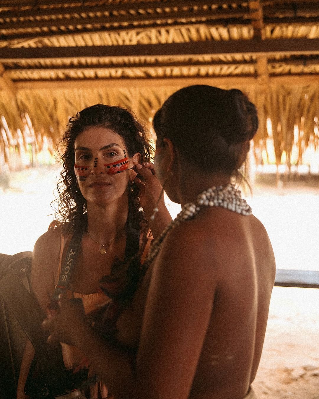 Eine indigene Frau trägt traditionelle rote und schwarze Gesichtsbemalung auf eine lächelnde Reisende in einer strohgedeckten Hütte im Juma Amazon Jungle Lodge auf