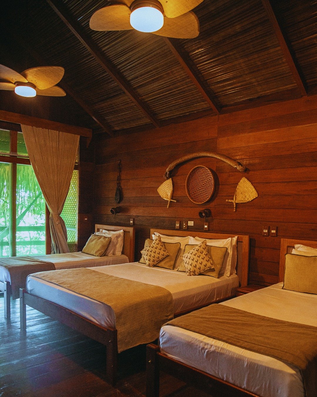 Dreibettzimmer in der Juma Amazon Jungle Lodge mit drei Holzbetten, gewebten Wanddekorationen, einem Strohdach mit Deckenventilatoren und Blick auf den Regenwald durch die Balkontür