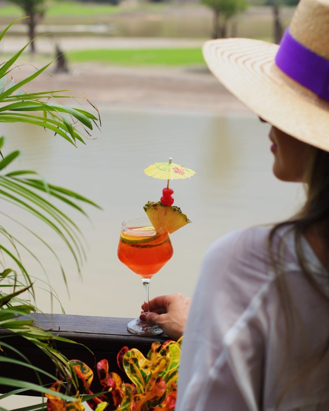A fruit-garnished cocktail, a straw hat, and the Amazon stretching out ahead. A woman in a wide-brimmed straw hat holds a vibrant orange tropical cocktail garnished with pineapple, cherry and a paper umbrella at Juma Amazon Jungle Lodge, with a tranquil waterway and lush greenery behind her