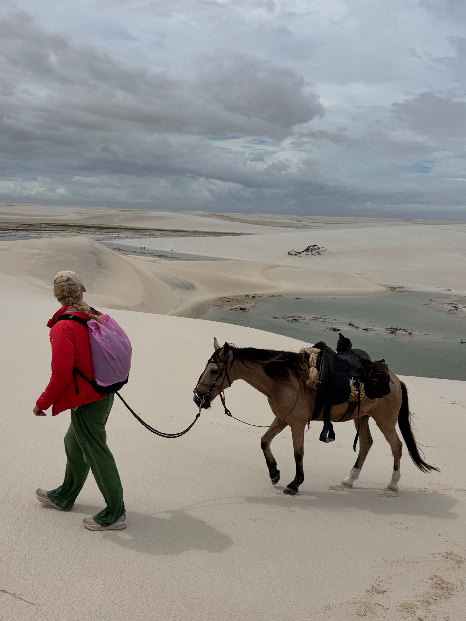 A traveler leads a horse on foot across the vast white dunes of Lençóis Maranhenses under a cloudy sky.