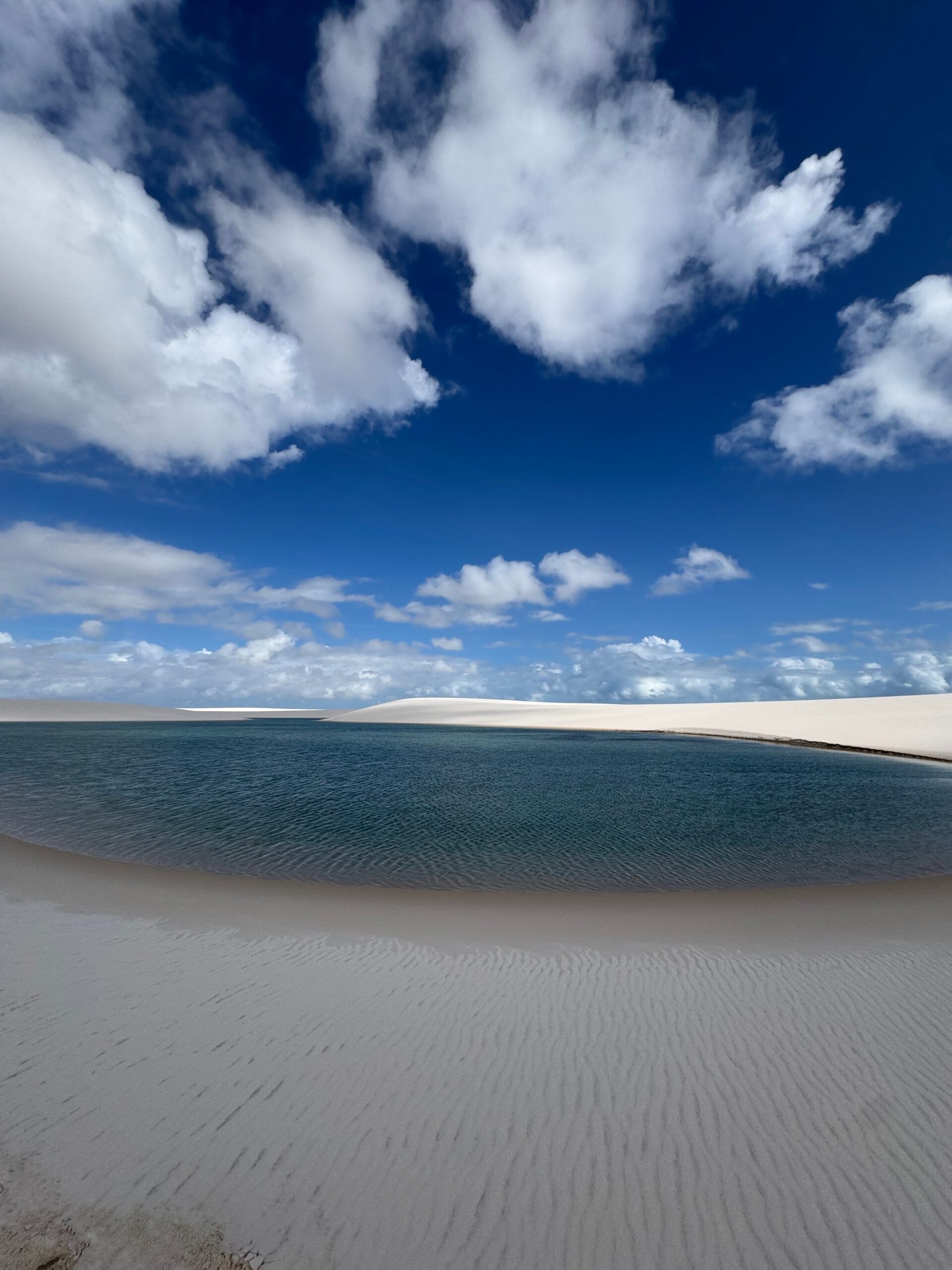 A turquoise rainwater lagoon nestled between white sand dunes under a bright blue sky in Lençóis Maranhenses.