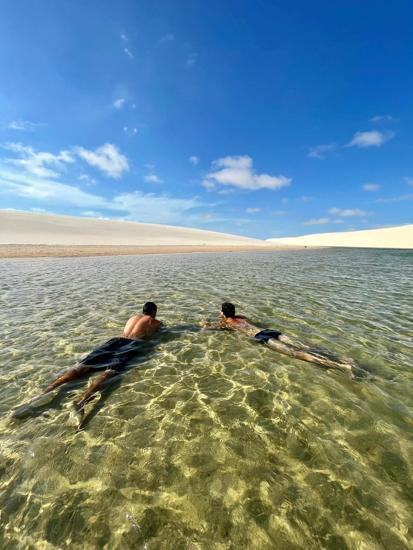 Cool, glass-clear water — the dunes can wait. Two swimmers gliding through crystal-clear shallow water in a Lençóis Maranhenses full itinerary lagoon, with sweeping white sand dunes stretching to the horizon under a vivid blue sky
