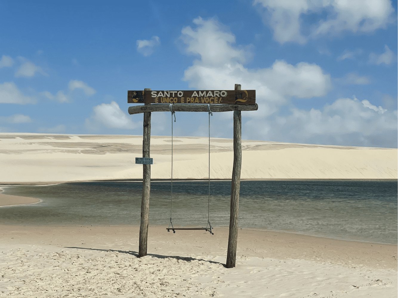 Santo Amaro's welcome: a rope swing, a lagoon, and nothing but dunes beyond. A rustic wooden swing and sign reading 'Santo Amaro É Único É Pra Você' framed by a turquoise lagoon and white sand dunes on the Lençóis Maranhenses full itinerary