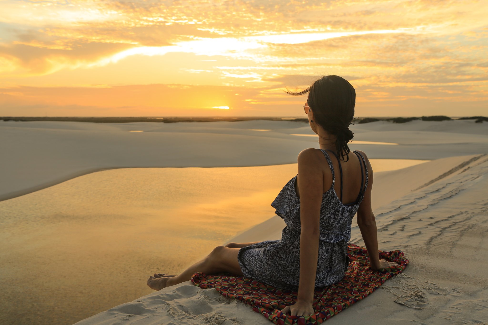 Bare feet, warm sand, and the whole sky on fire. Photo: Brunomsbarreto A woman sits on a colorful blanket atop a white sand dune, watching a vivid golden sunset spread across the sky and reflect off still water in the Lençóis Maranhenses full itinerary landscape below