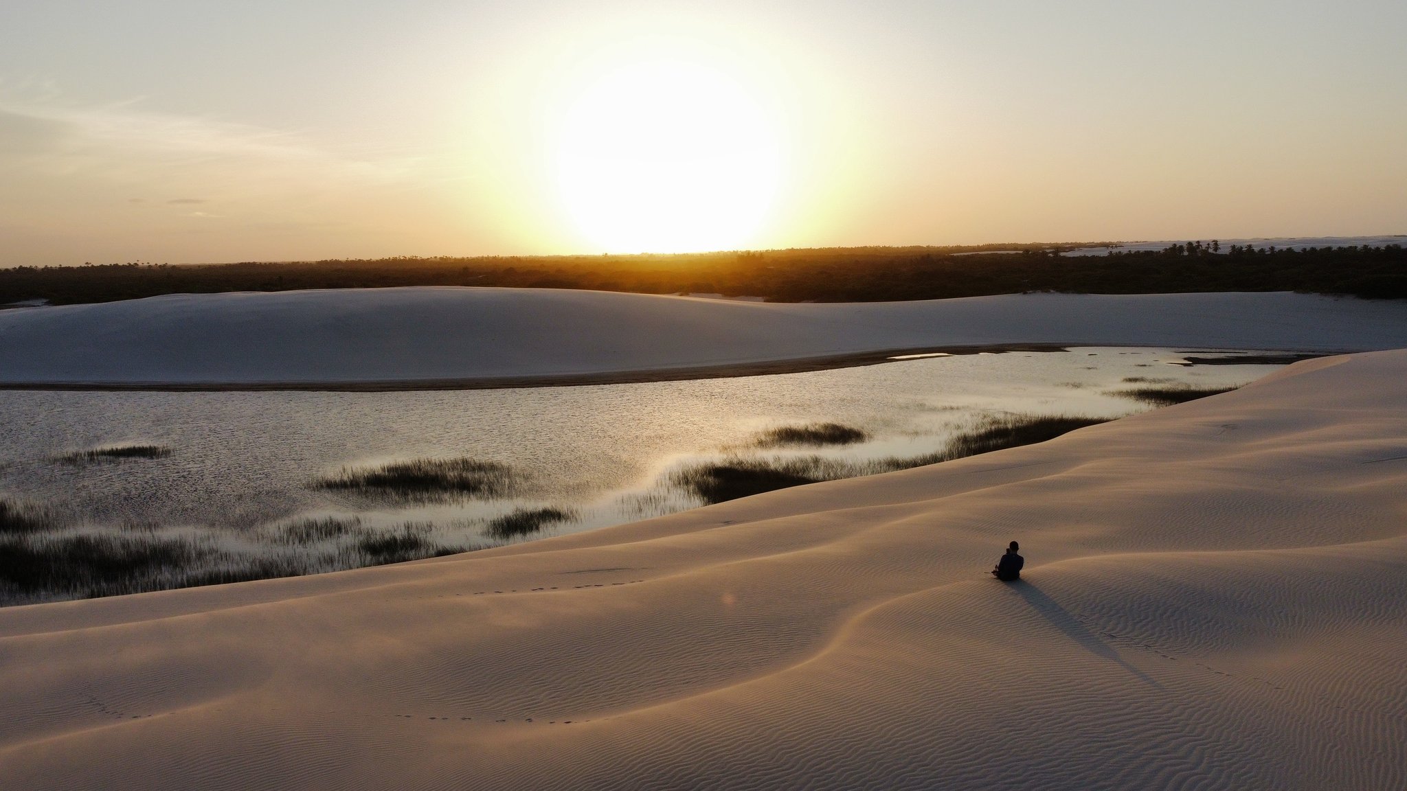 Still on the dune's edge, the sun melting into the horizon — solitude at its most golden. A lone figure sits on rippled white sand dunes overlooking a shallow lagoon fringed with grass at golden sunset, part of the Lençóis Maranhenses full itinerary experience